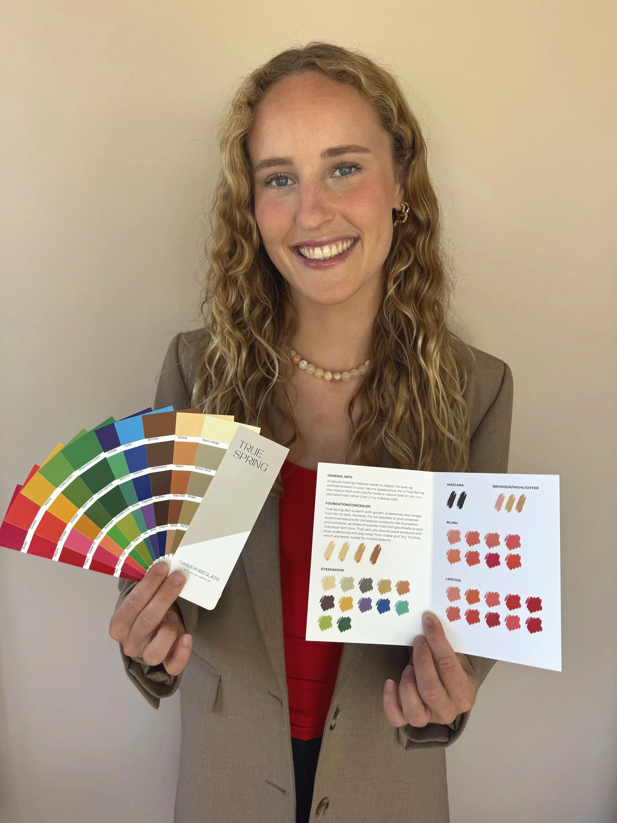 Kvinde med krøllet hår og smilende ansigt holder farvekort og produktbeskrivelse om makeup, herunder farver til foundation, øjenskygge, læbestift og blush. Baggrunden er neutral.