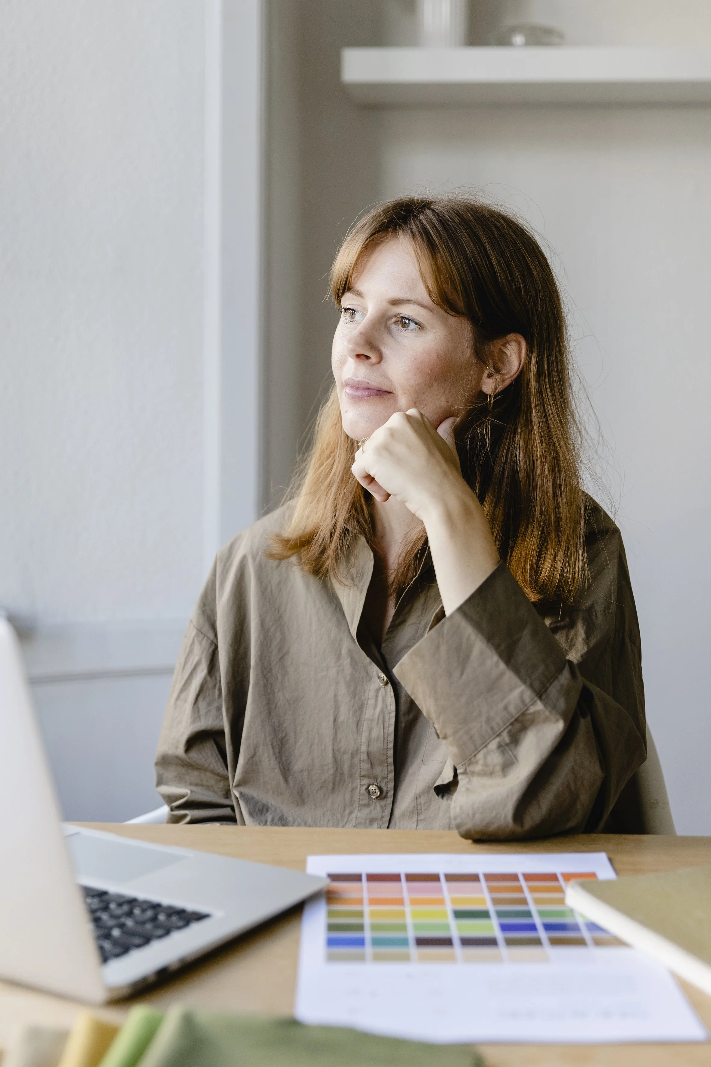 Kvindelig person med rød hår, der sidder ved et bord, omgivet af farvekort, en bærbar computer og papirer. Personen har en tænksom udtryks, hviler sit ansigt på sin hånd og ser ud af vinduet.