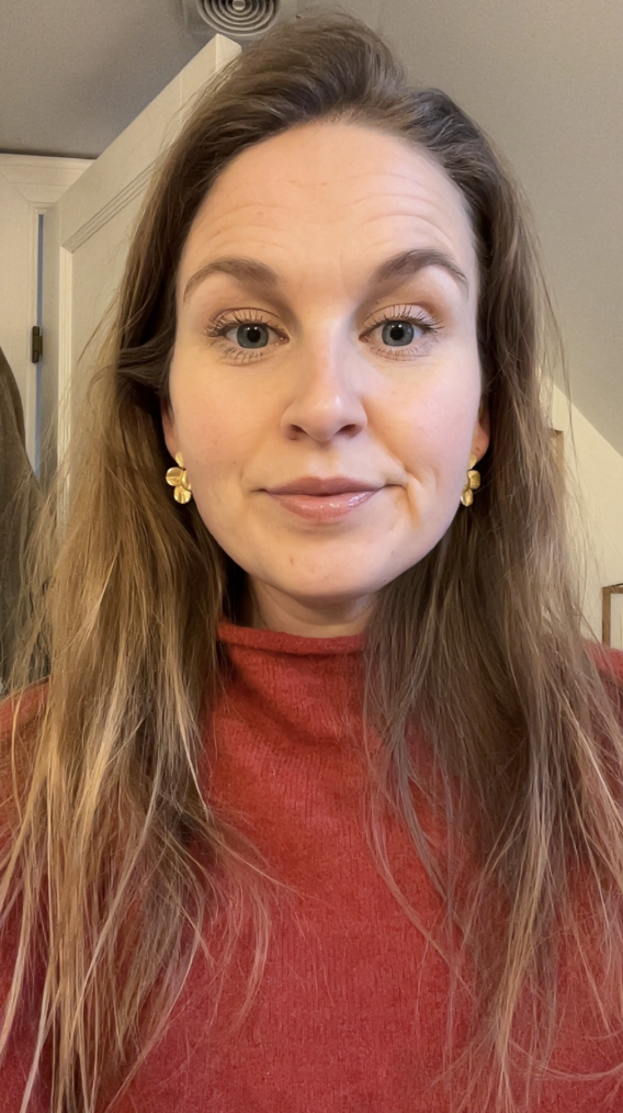 Et close-up af en kvinde med langt, lyst hår, iført en rød skjorte og guld øreringe, i et indendørs miljø.