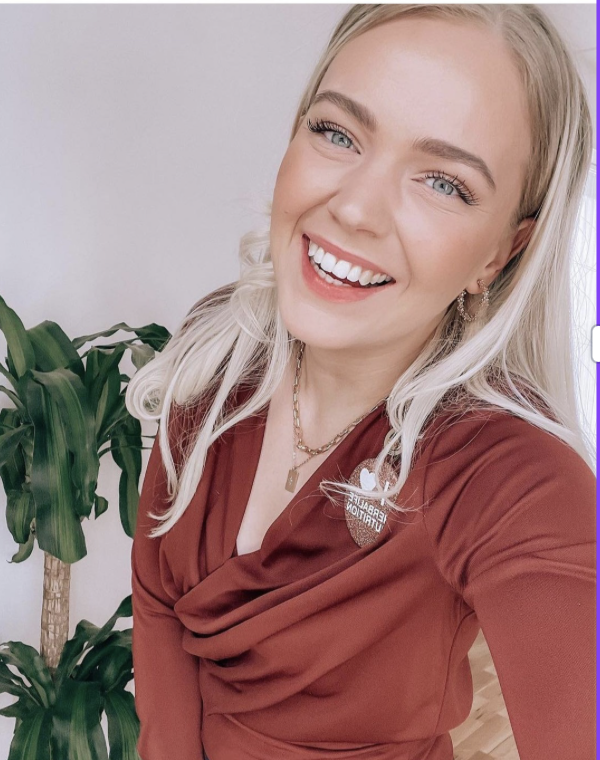 Smilende kvinde med blond hår iført en rød kjole, stående ved en stor grøn plante.