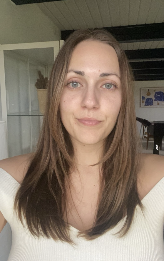 Nærbillede af en ung kvinde med langt brunt hår, blå øjne og let makeup, smilede let foran et hjemmemiljø.