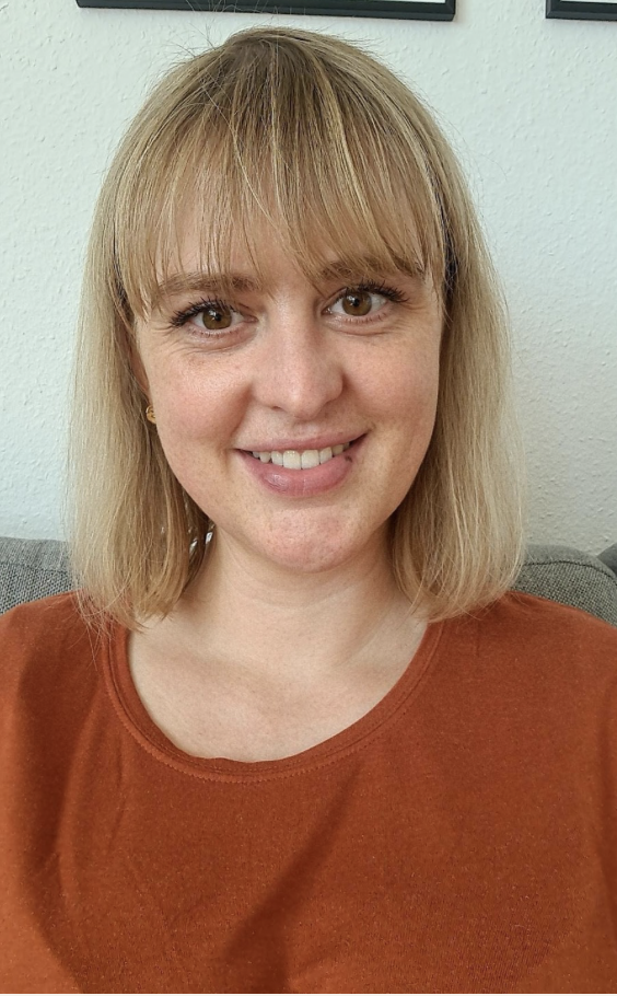 En smilende kvinde med blond hår og brune øjne, iført en rustfarvet T-shirt, sidder på en sofa med en hvid væg bag sig.