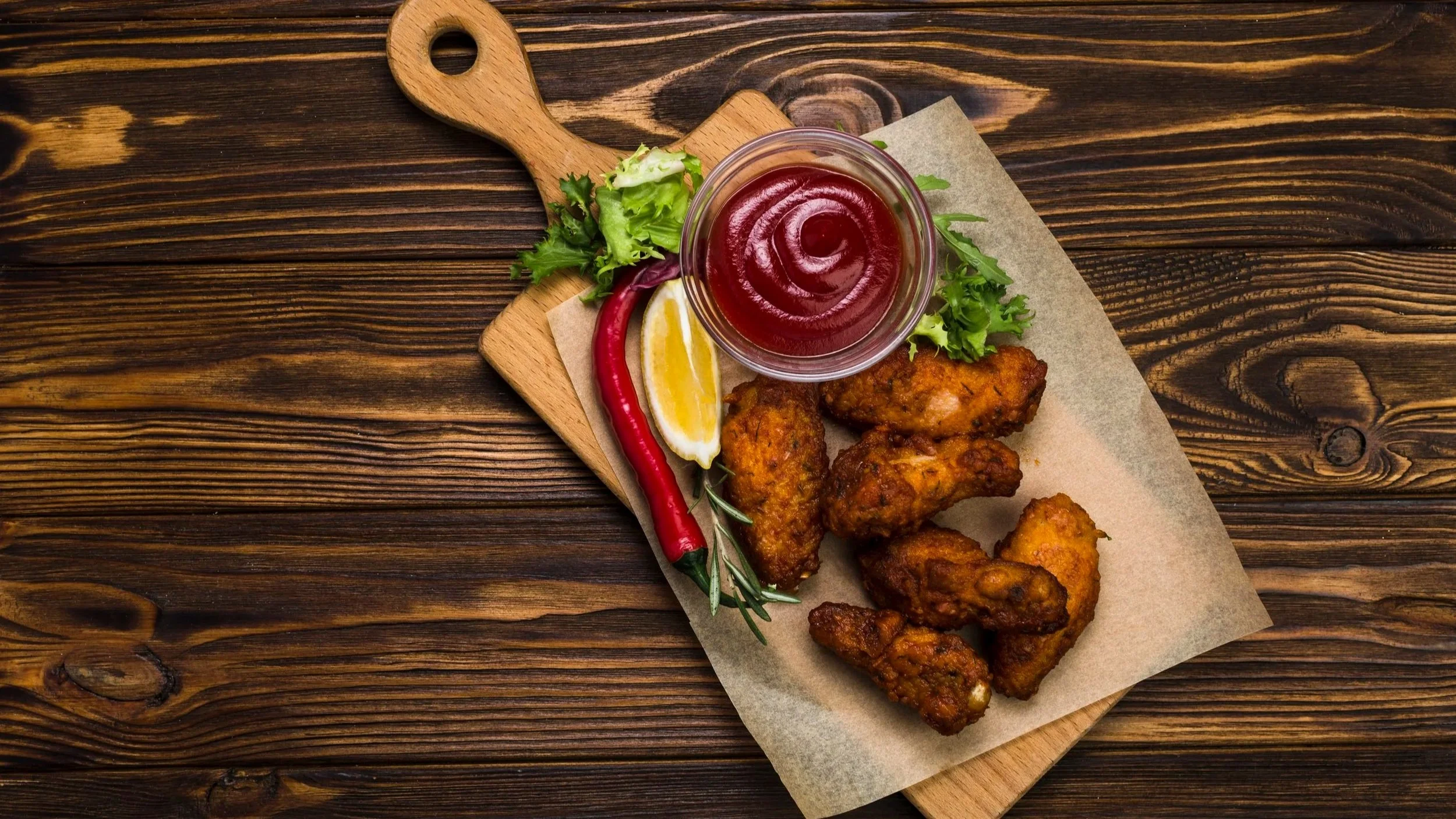 Fried chicken drumsticks served with a side of salad, lemon wedge, chili pepper, barbecue sauce, and fresh herbs on a wooden serving board.