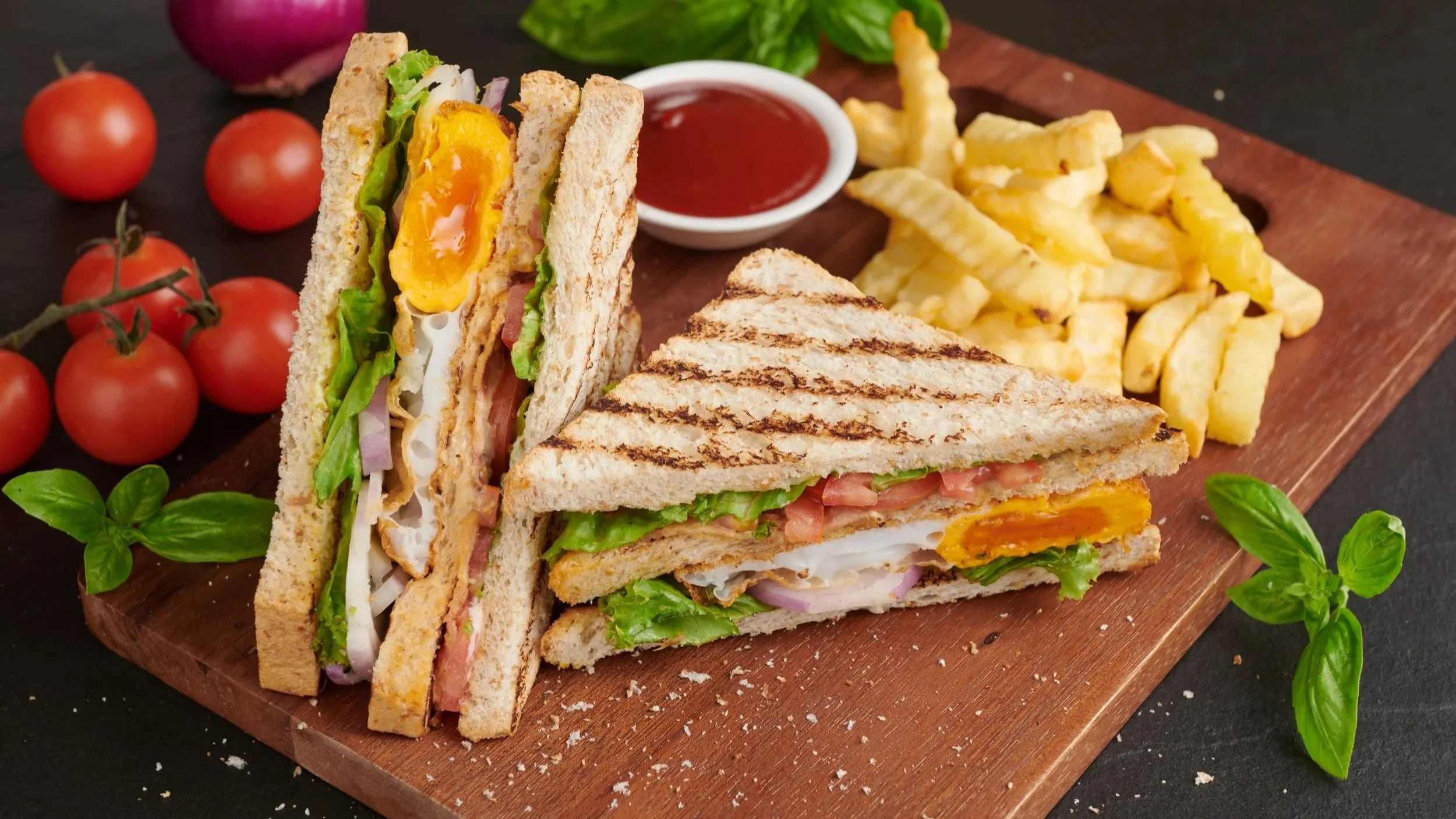 A wooden serving board with a sandwich cut into triangles, containing fried egg, lettuce, tomato, ham, and cheese, with fresh cherry tomatoes, basil leaves, and a small bowl of ketchup on the side.