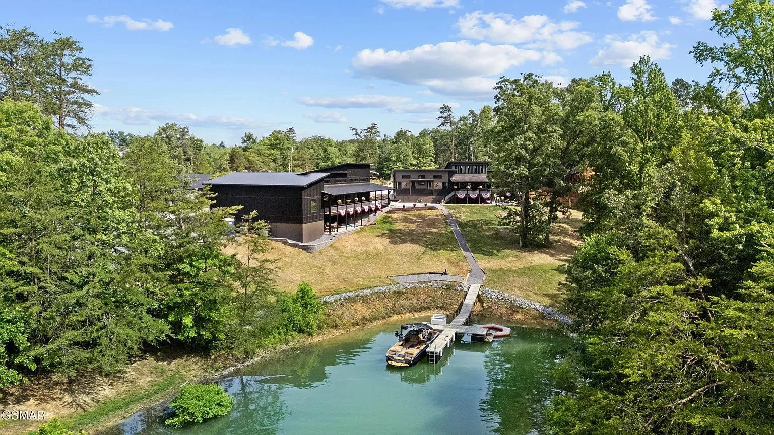A lakeside property with a dock and boat, surrounded by lush green trees and a grassy hill with a modern black building, under a partly cloudy blue sky.