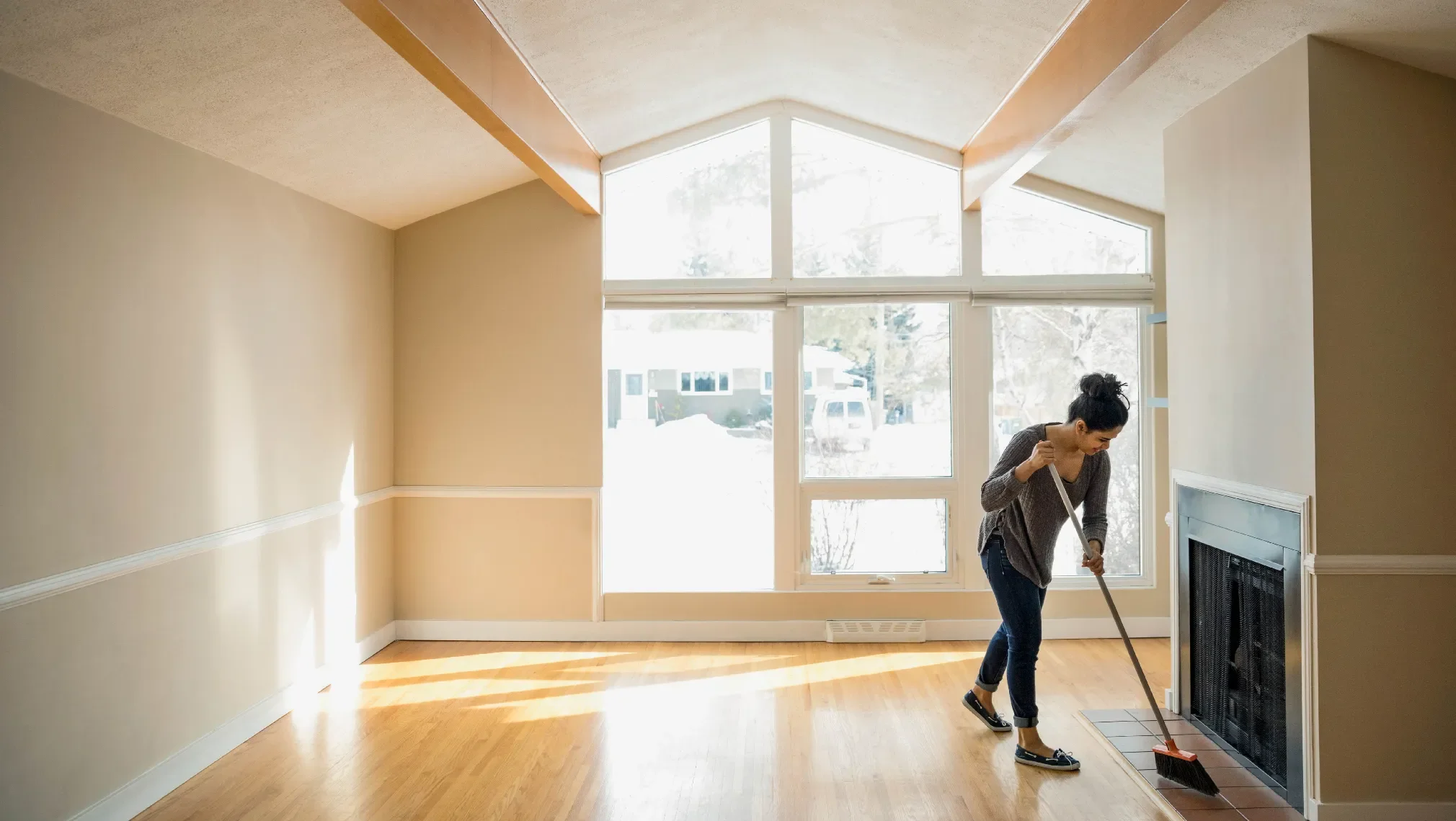A woman cleaning a living room with large windows, a fireplace, and hardwood floors.