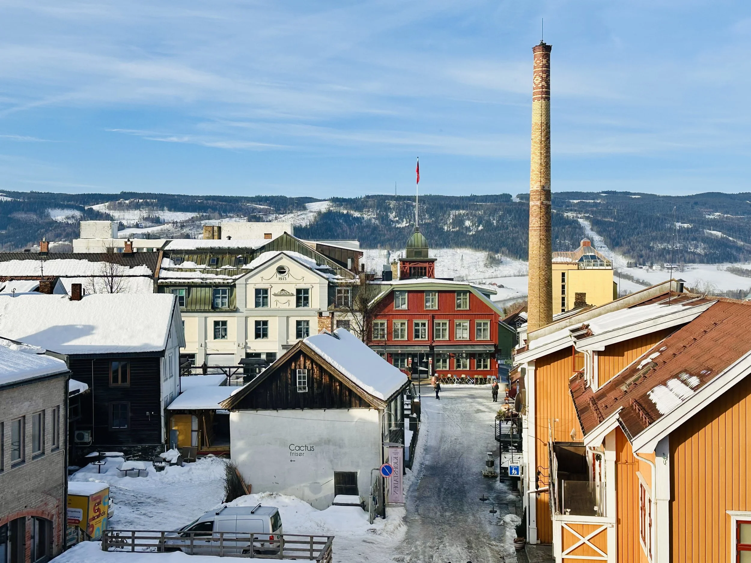 Snow-covered buildings and streets with a tall chimney, hills in the background, and a partly cloudy sky.