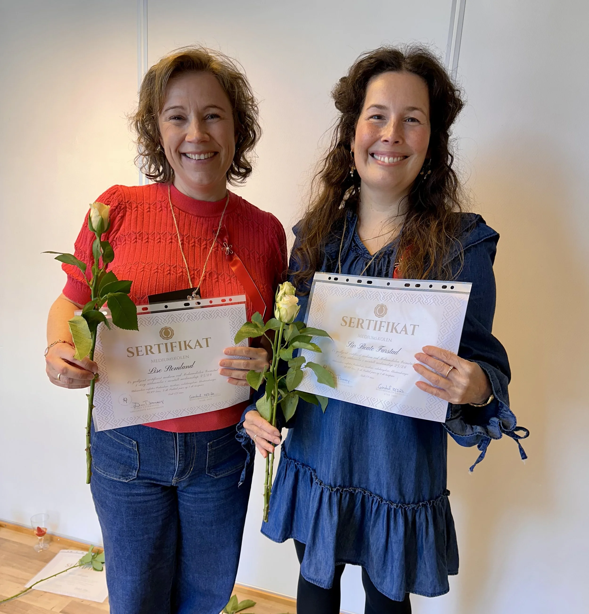 Two women standing side by side, smiling, holding certificates and white roses. One woman has short curly hair and is wearing a red sweater, while the other has long curly hair and is wearing a dark blue dress. They are in a room with a plain cream wall background, and there is a glass of red drink on the floor near them.