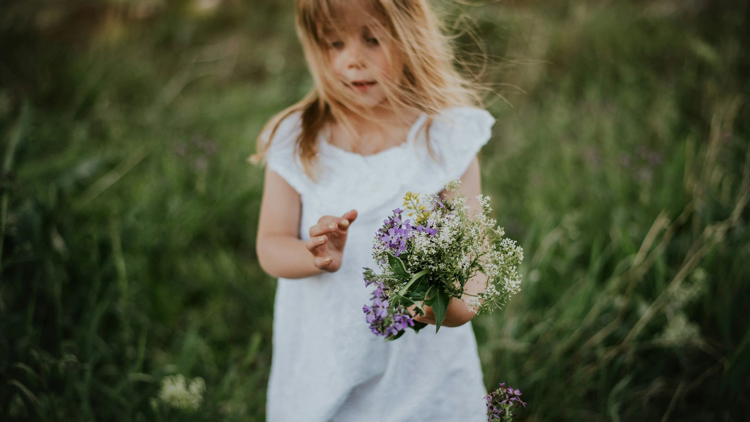 A young girl with reddish hair wearing a white dress, standing outdoors in a grassy area, holding a colorful bouquet of wildflowers. She is looking downward at the flowers.