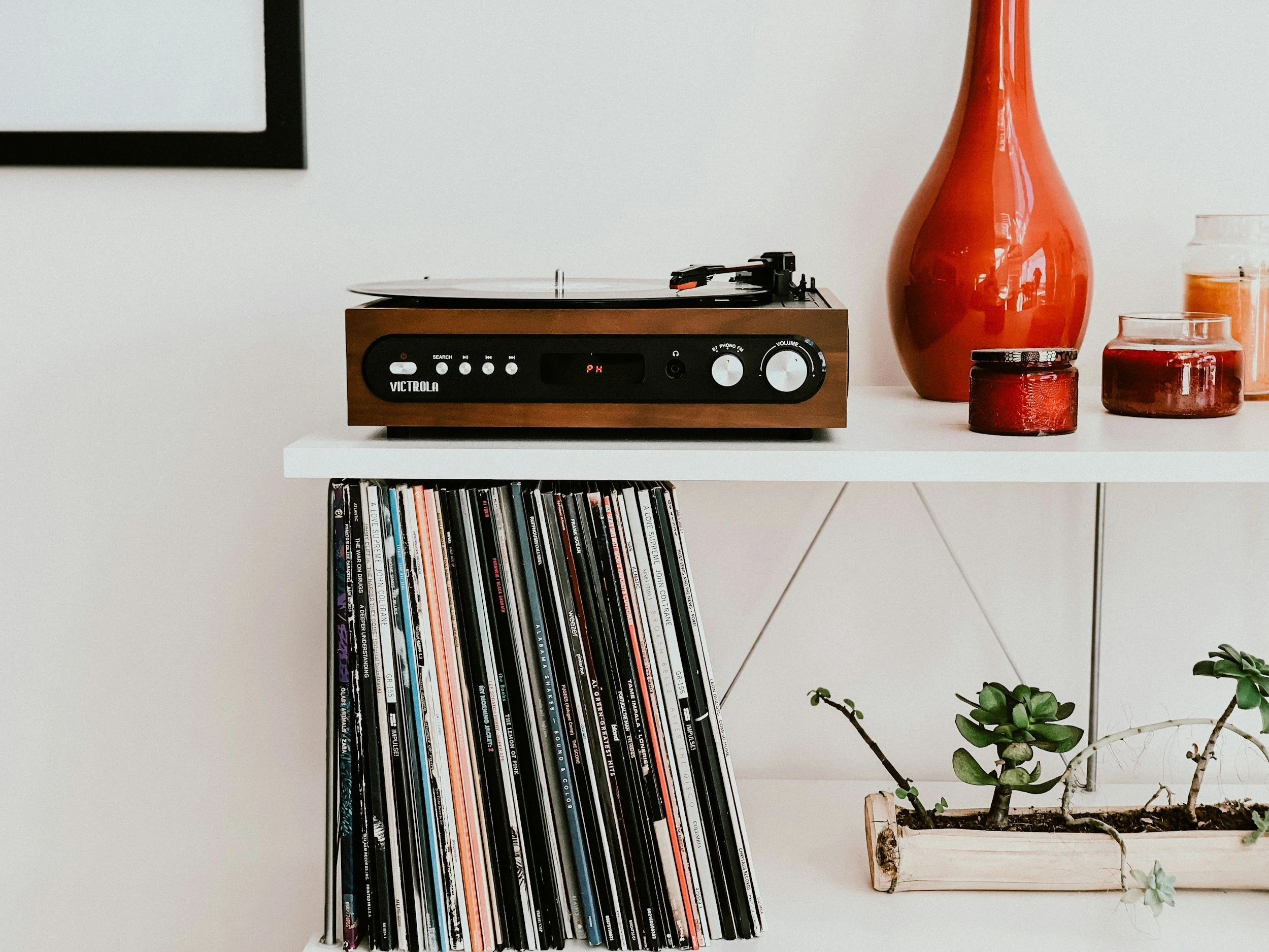 A white shelf holding a record player, vinyl records, a large orange vase, a glass jar with orange contents, a small candle in a jar, and a potted succulent plant.