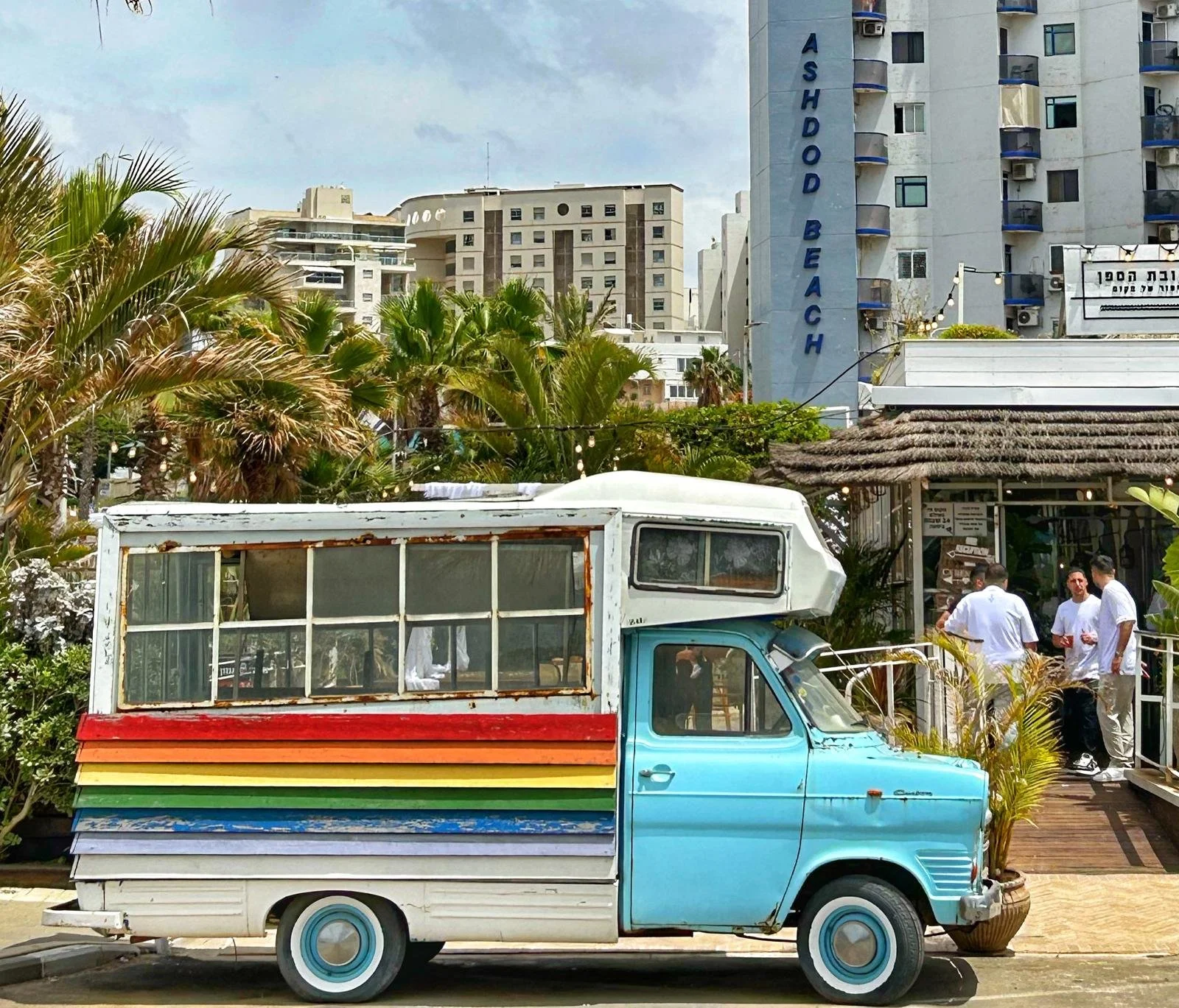 A colorful small vintage truck with rainbow stripes parked near a restaurant called 'Ashdod Beach' in a city with high-rise buildings in the background. There are some people standing near the restaurant entrance.