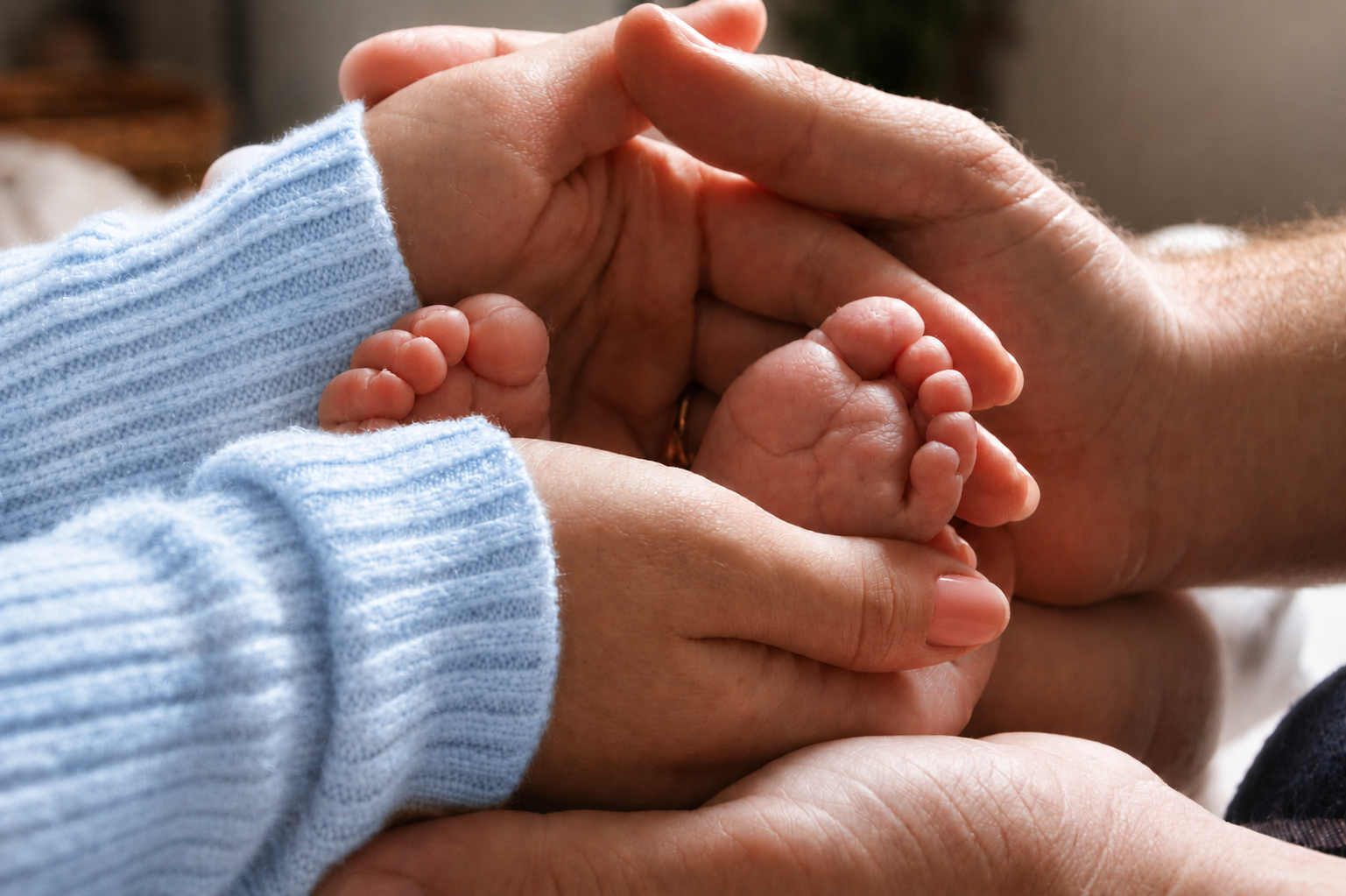 Close-up of parents' hands cradling a baby's tiny feet, with one parent wearing a blue sweater.