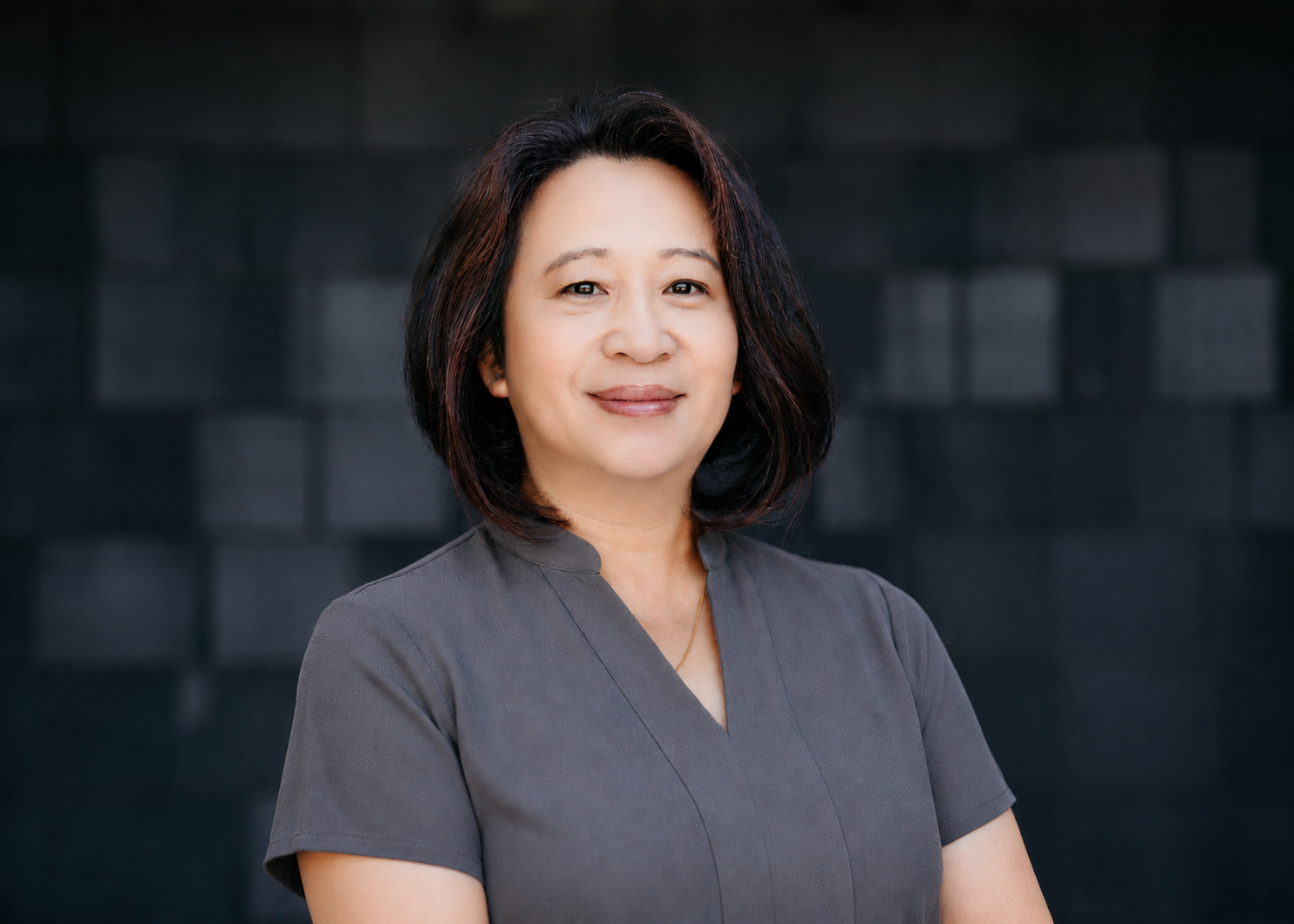 A woman with shoulder-length black hair, wearing a dark gray top, smiling softly against a black brick wall background.