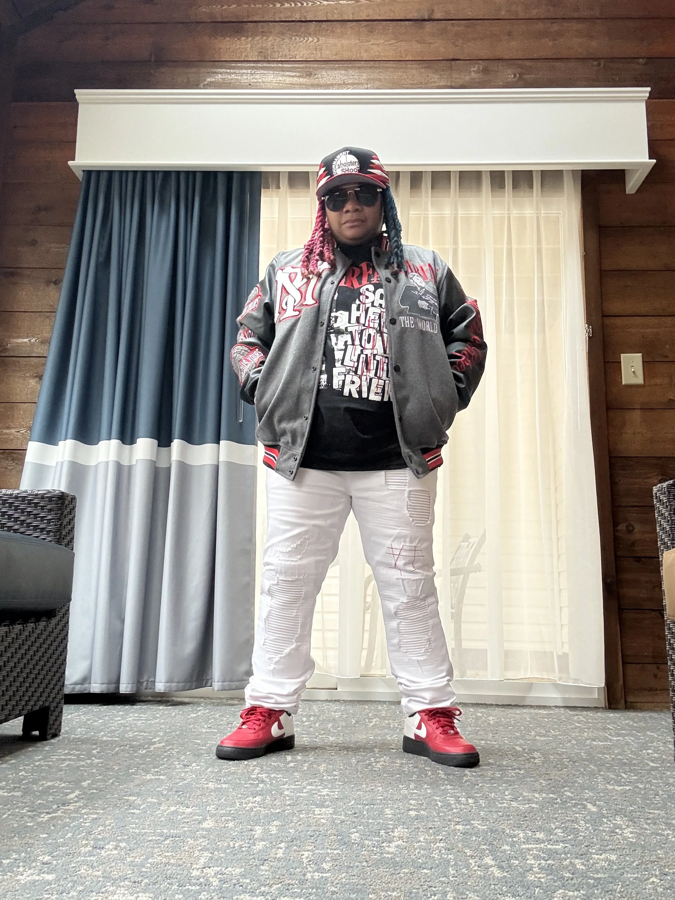 Person standing indoors in front of a glass door with curtains, wearing a gray and red varsity jacket, a black graphic T-shirt, white distressed jeans, red and white sneakers, sunglasses, and a baseball cap with a red, white, and black pattern.