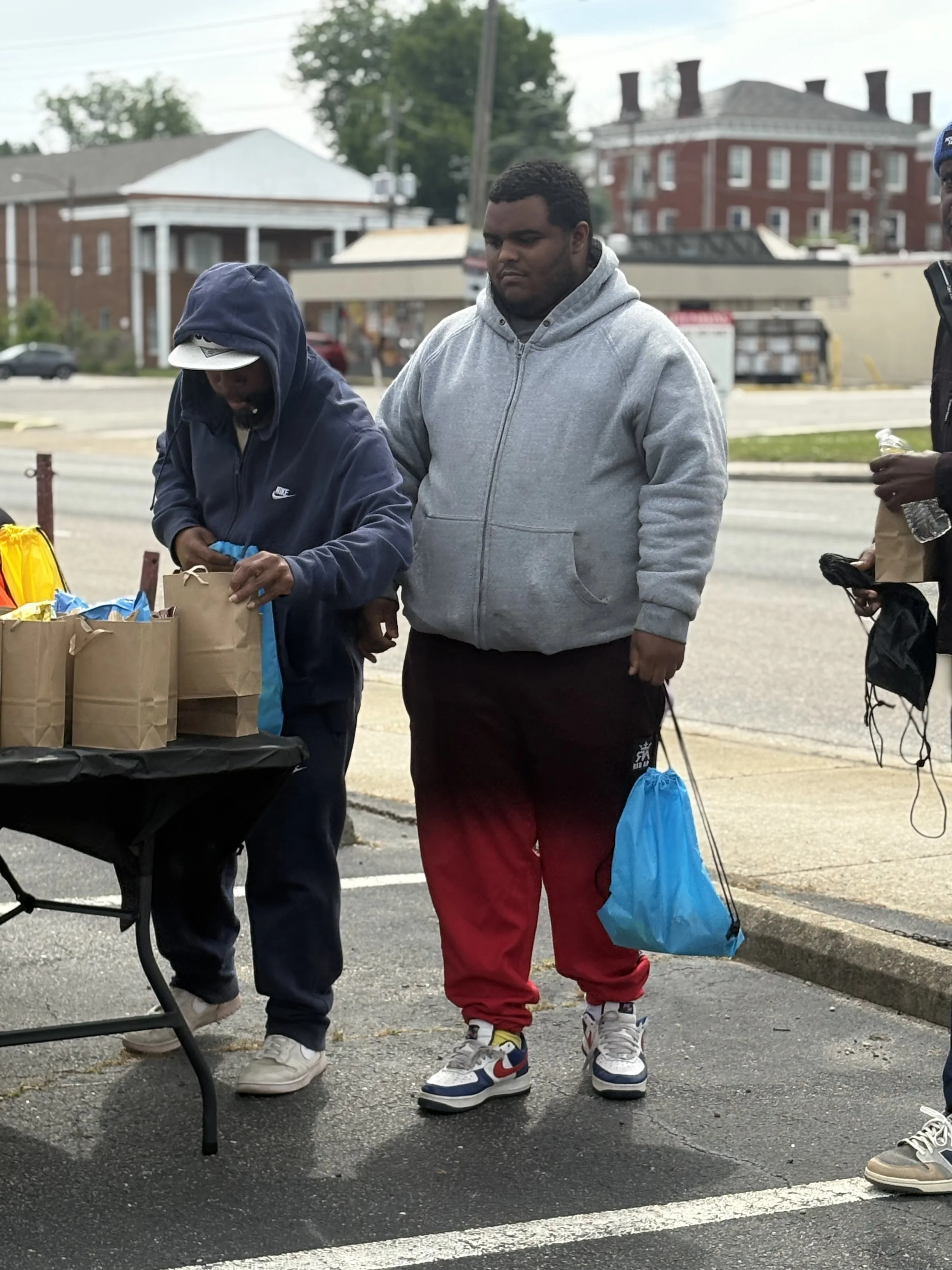 Three young men standing outdoors near a table with brown paper bags, with buildings and trees in the background. One man wears a hoodie and cap, another wears a grey hoodie, and the third is partially visible holding a water bottle.