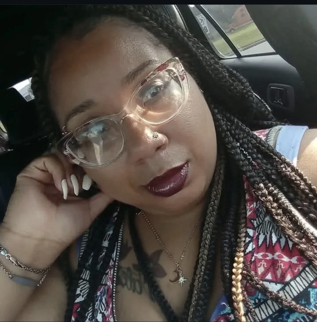 A woman with dark lipstick and clear glasses taking a selfie inside a car, with a blurred landscape of trees and houses outside the window.