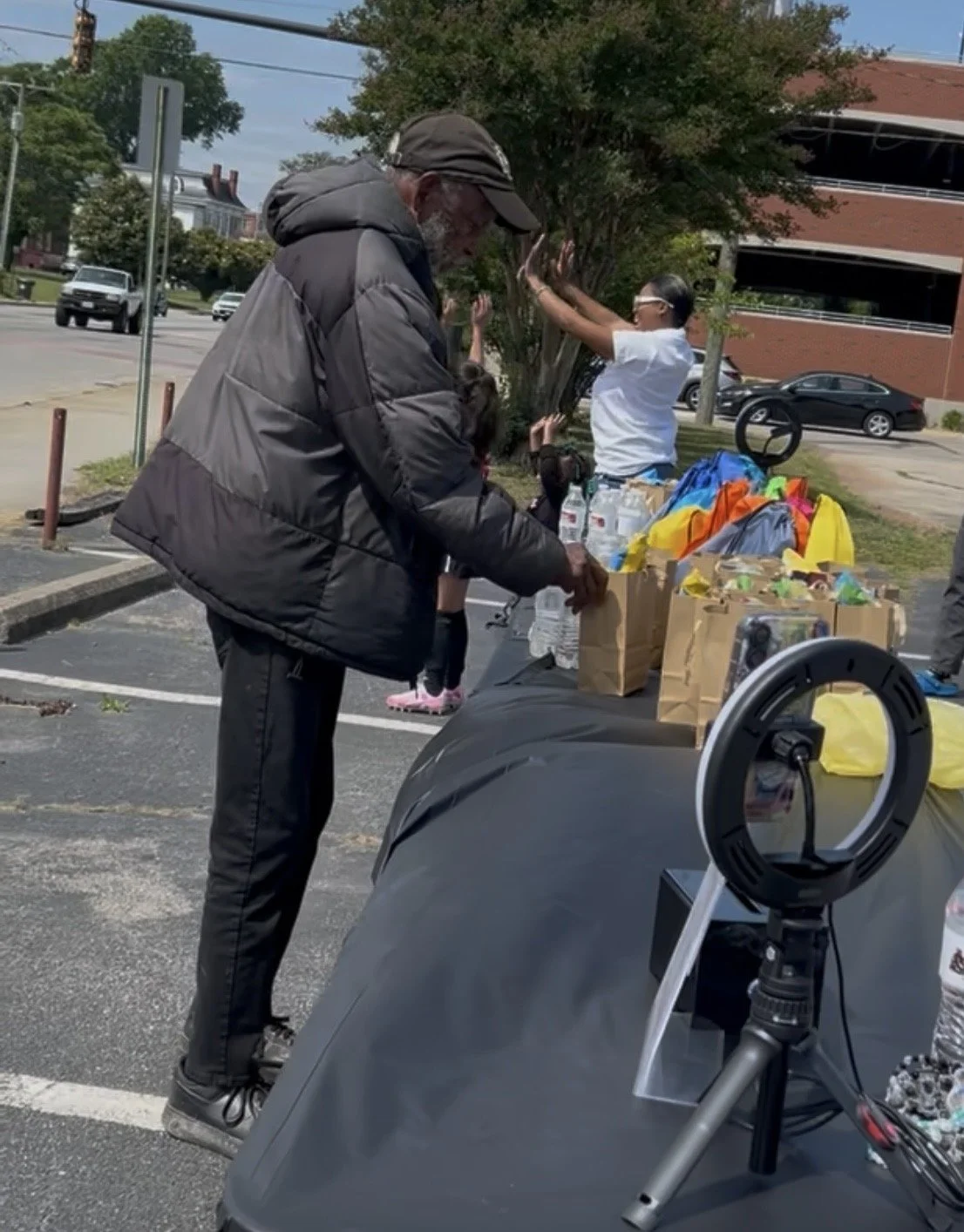 A man in a black jacket at outdoor charity event, placing items into a paper bag on a table. A woman with sunglasses stands behind the table, reaching for items, with colorful bags and bottled water on the table. A ring light and camera are visible in the foreground. Cars and a brick building are in the background.