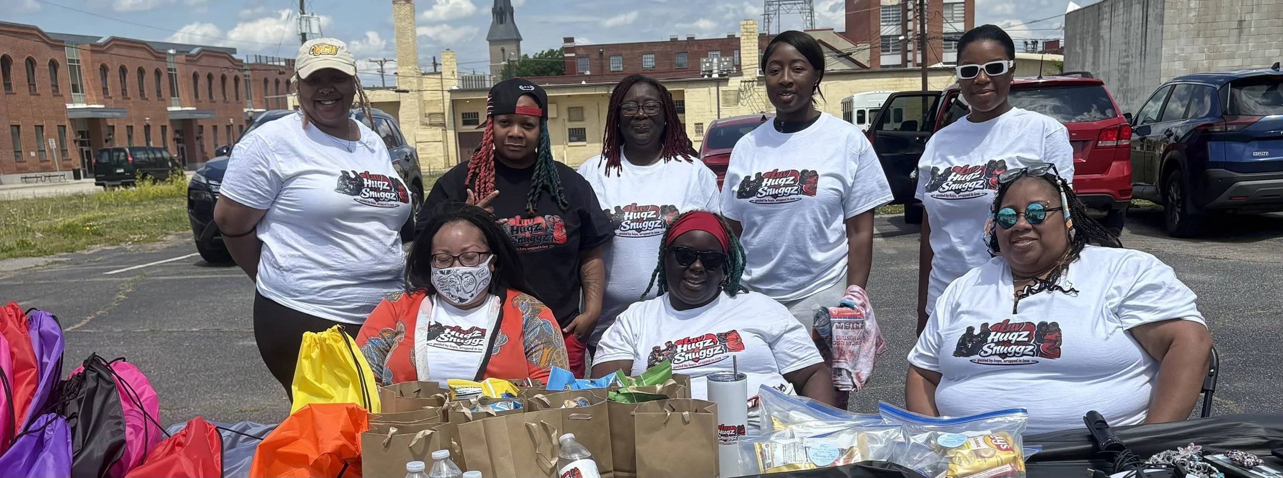 Group of women posing outdoors behind a table with food and supplies, some wearing shirts with the logo 'Hugz & Snugz'.