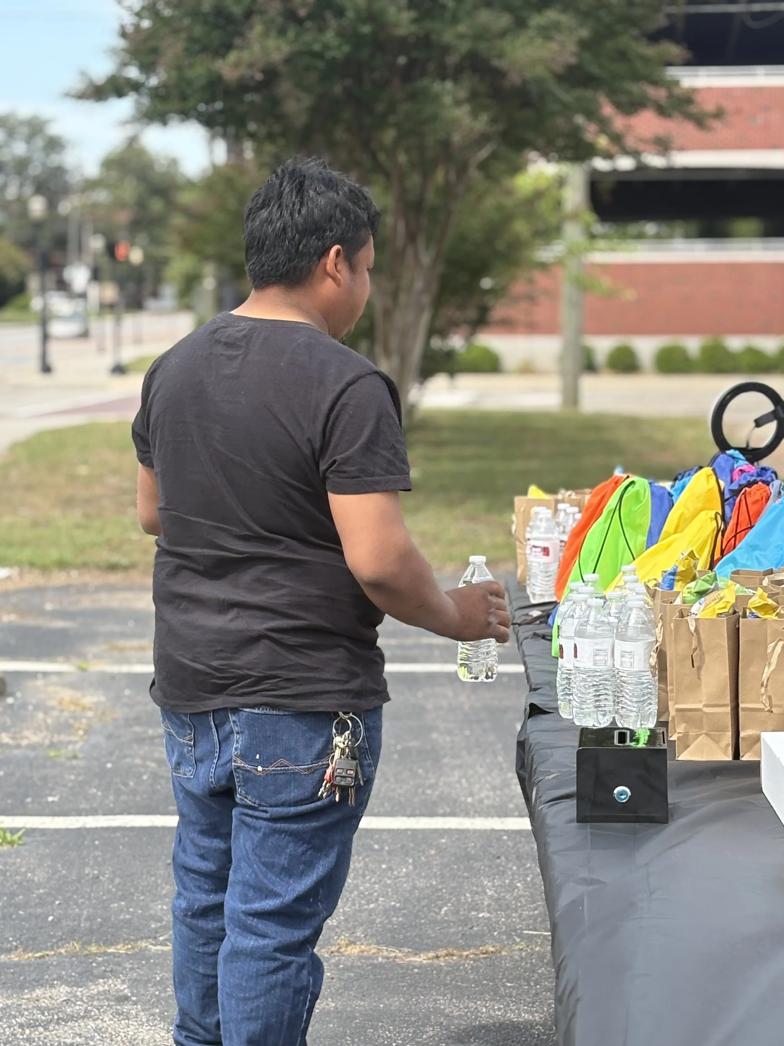 A man with a keychain hanging from his jeans standing outdoors at a table with bottled water and colorful bags, possibly volunteering or attending an event.