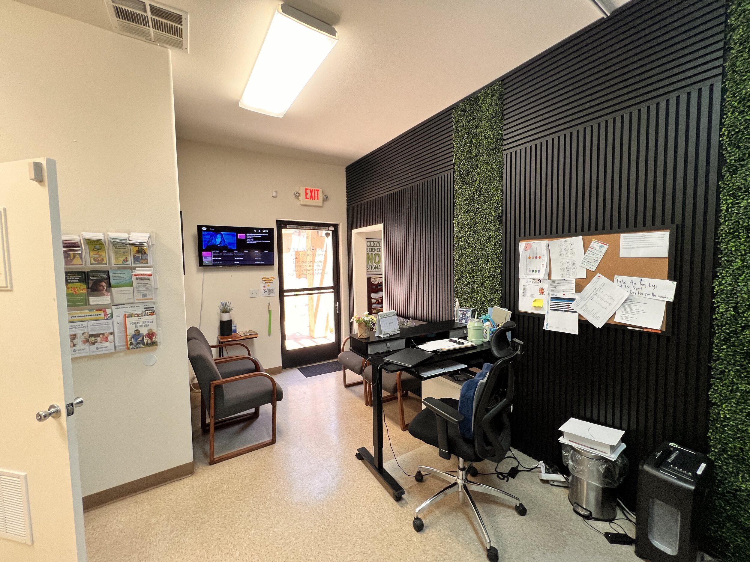 An office reception area with chairs, a desk with a computer, a bulletin board, and green wall decor.