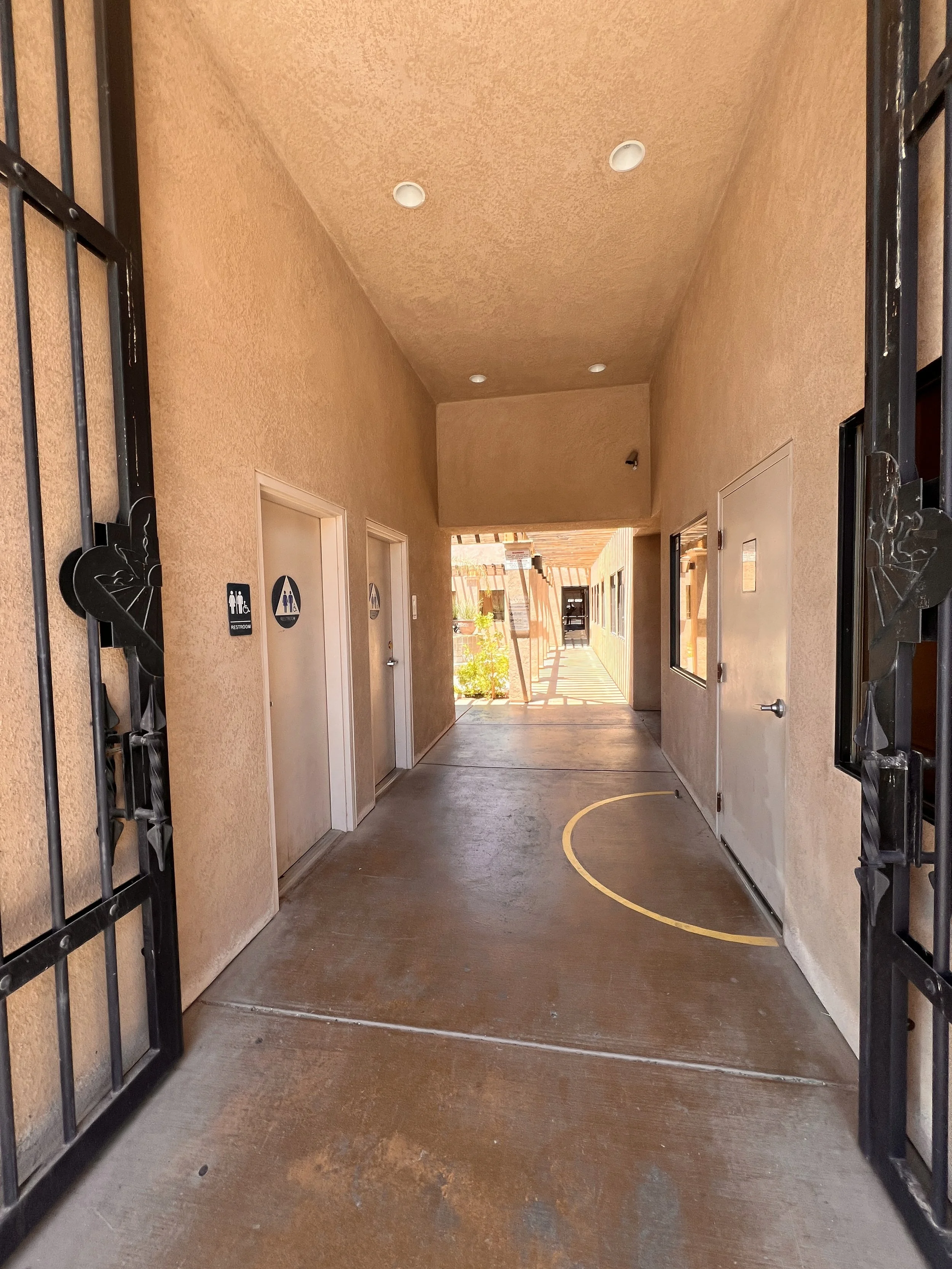 A corridor with beige stucco walls, several restroom doors marked male and female, ceiling lights, and a shaded outdoor area at the end of the corridor.
