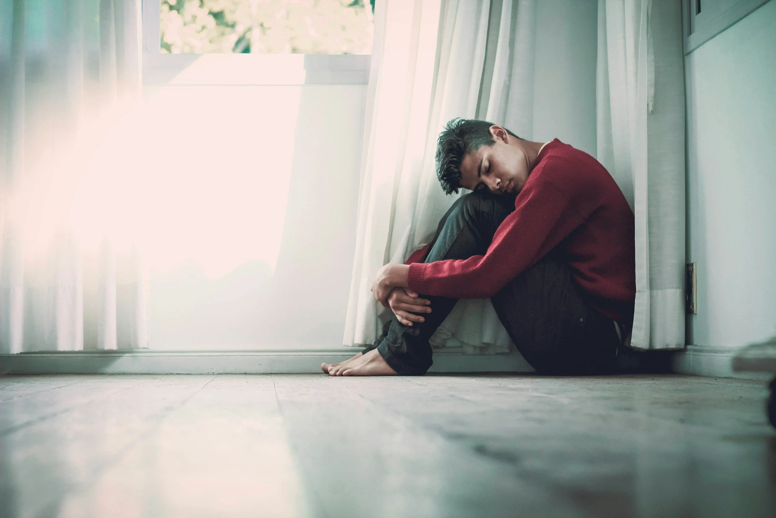 Young man sitting on the floor by a window with white curtains, with his knees drawn up and arms wrapped around his legs, looking downward with a somber expression.