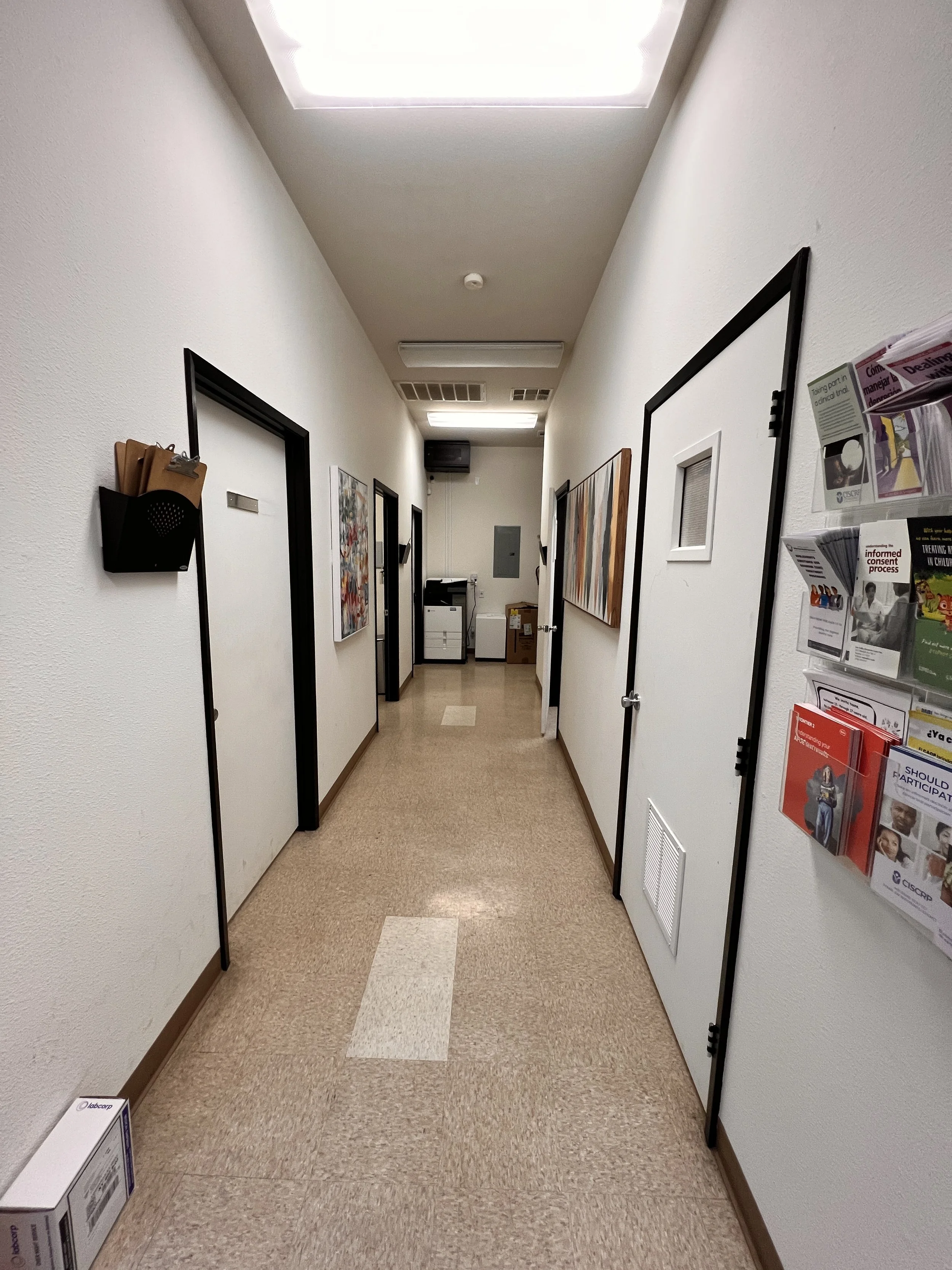 A hallway in an office or medical building with white walls, brown trim, vinyl tiled floor, revealing multiple closed doors and framed artwork hanging on the walls, a brochure rack on the right, and a printer and boxes at the end of the hallway.
