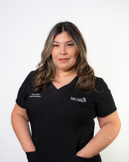 Woman with wavy hair wearing a black medical scrub top with the Sun Valley logo, standing against a white background.