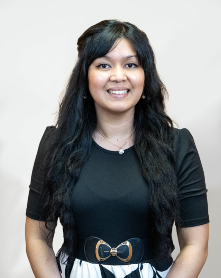 A woman with long, wavy black hair and a broad smile, wearing a black short-sleeve top with a bow belt, standing against a plain light background.