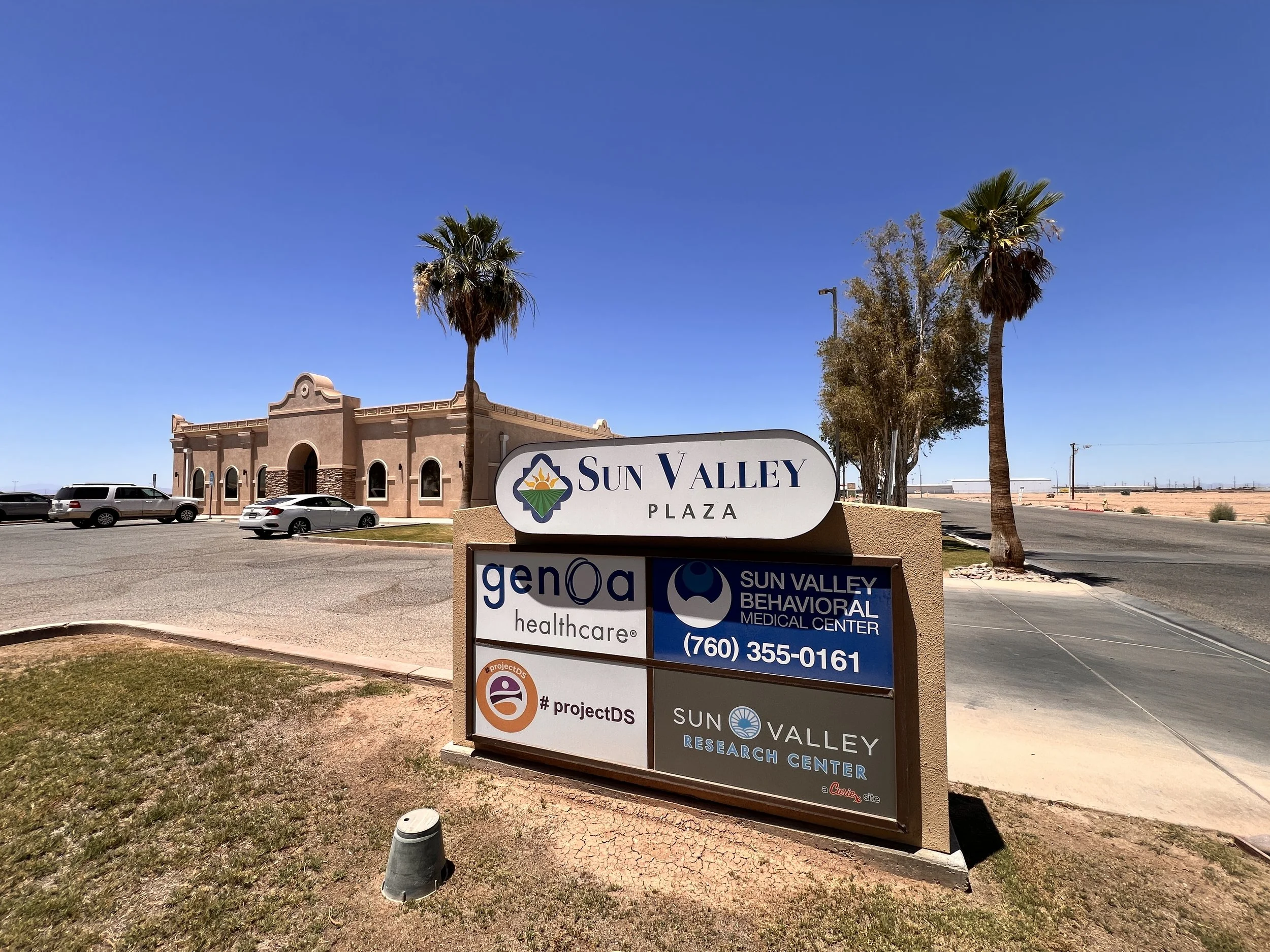 A large sign with multiple business names and logos in front of a beige building with arched windows, palm trees, and parked cars in a parking lot, under a clear blue sky.