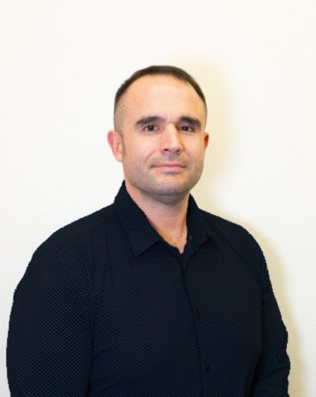 A man with short dark hair and light skin posing in front of a plain off-white wall, wearing a black collared shirt.