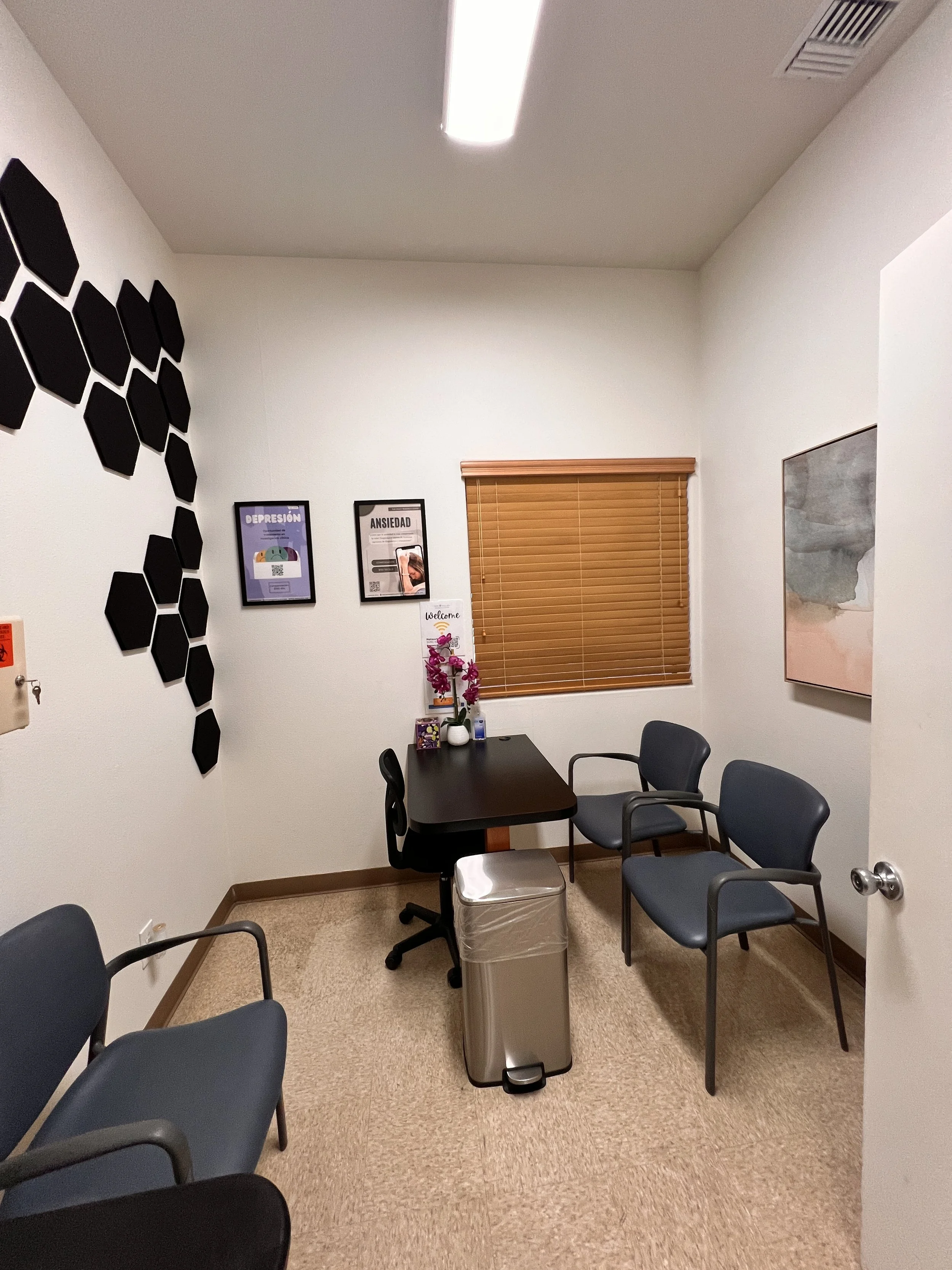 A small conference room with a black table, several blue chairs, and a stainless steel trash can. The room has beige carpet, white walls with framed posters, a window with wooden blinds, and a painting on the wall.