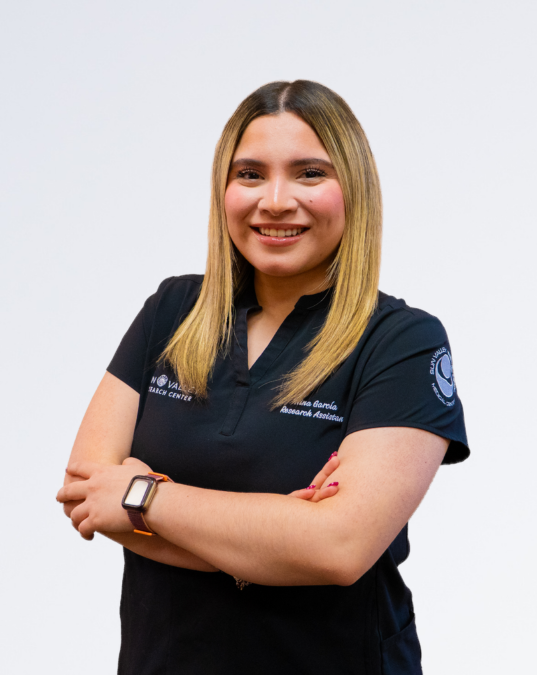 A young woman with shoulder-length blonde hair, smiling, wearing a black polo shirt with logos and text, standing against a plain white background.