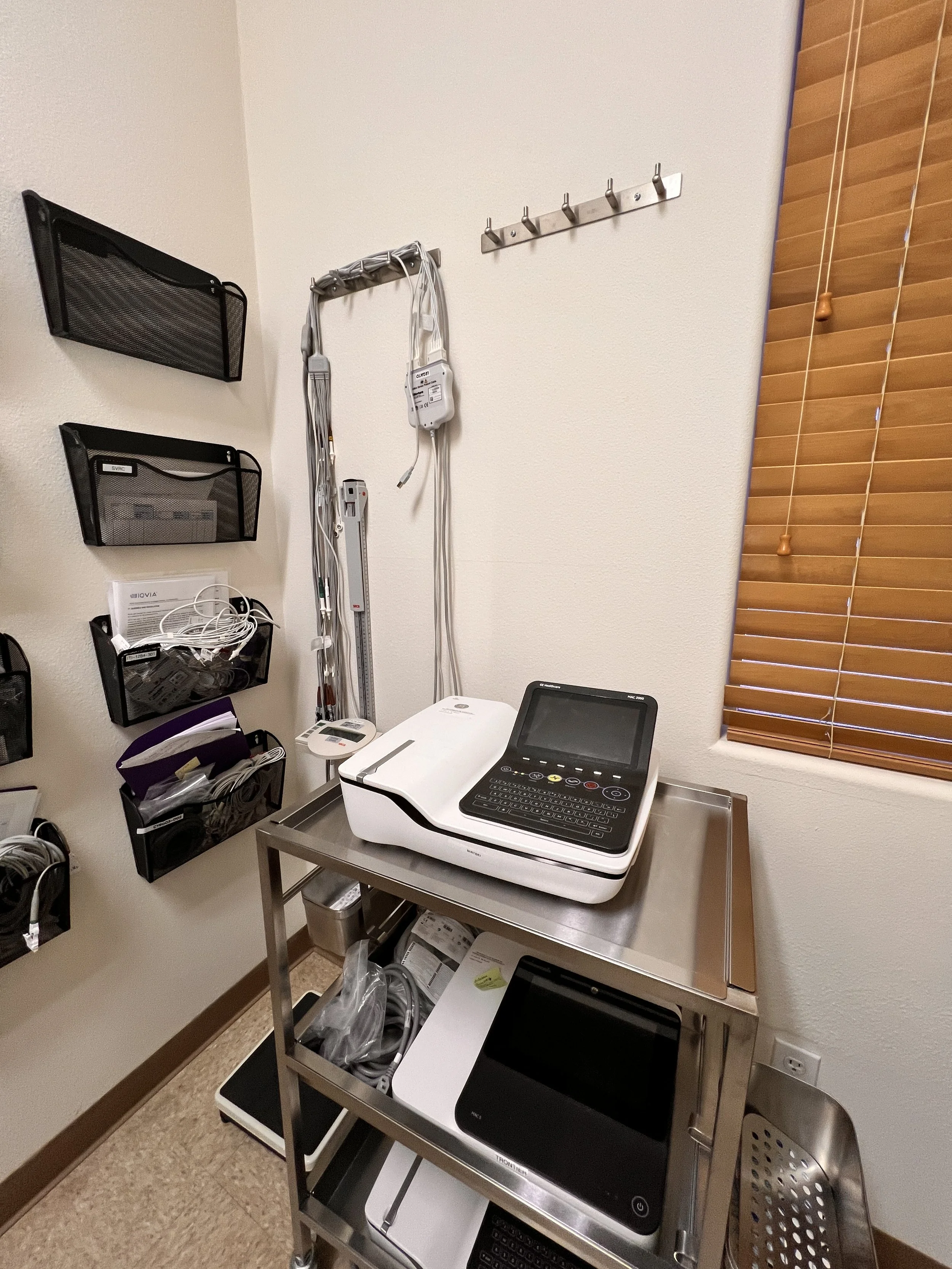 Medical equipment area with a stainless steel table holding an X-ray machine and a tablet, wall-mounted cable organizer, markers, and a window with wooden blinds.