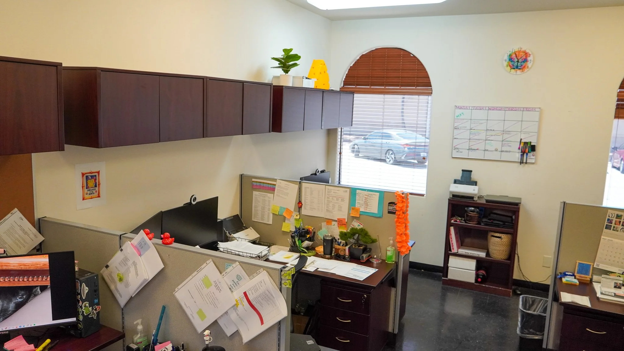 Office cubicles with computer monitors, paperwork, and office supplies, with a wall featuring storage cabinets, a window with blinds, and a whiteboard.