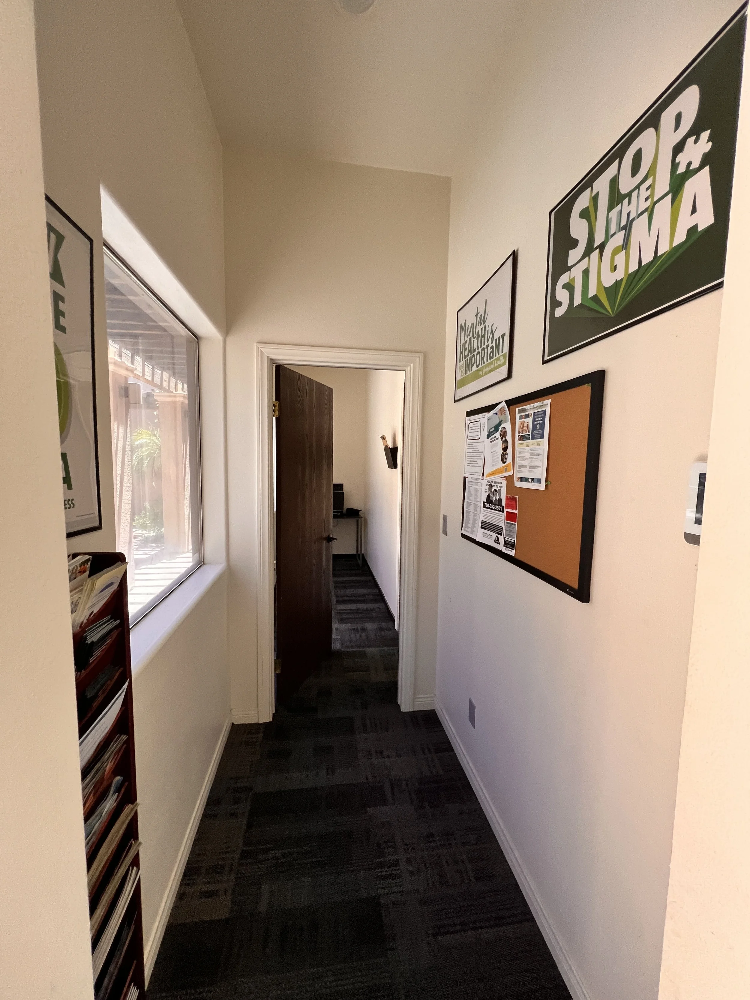 A hallway with a window on the left, a door at the end open revealing another room, and framed posters on the wall including a large one that says "STOP the STIGMA."