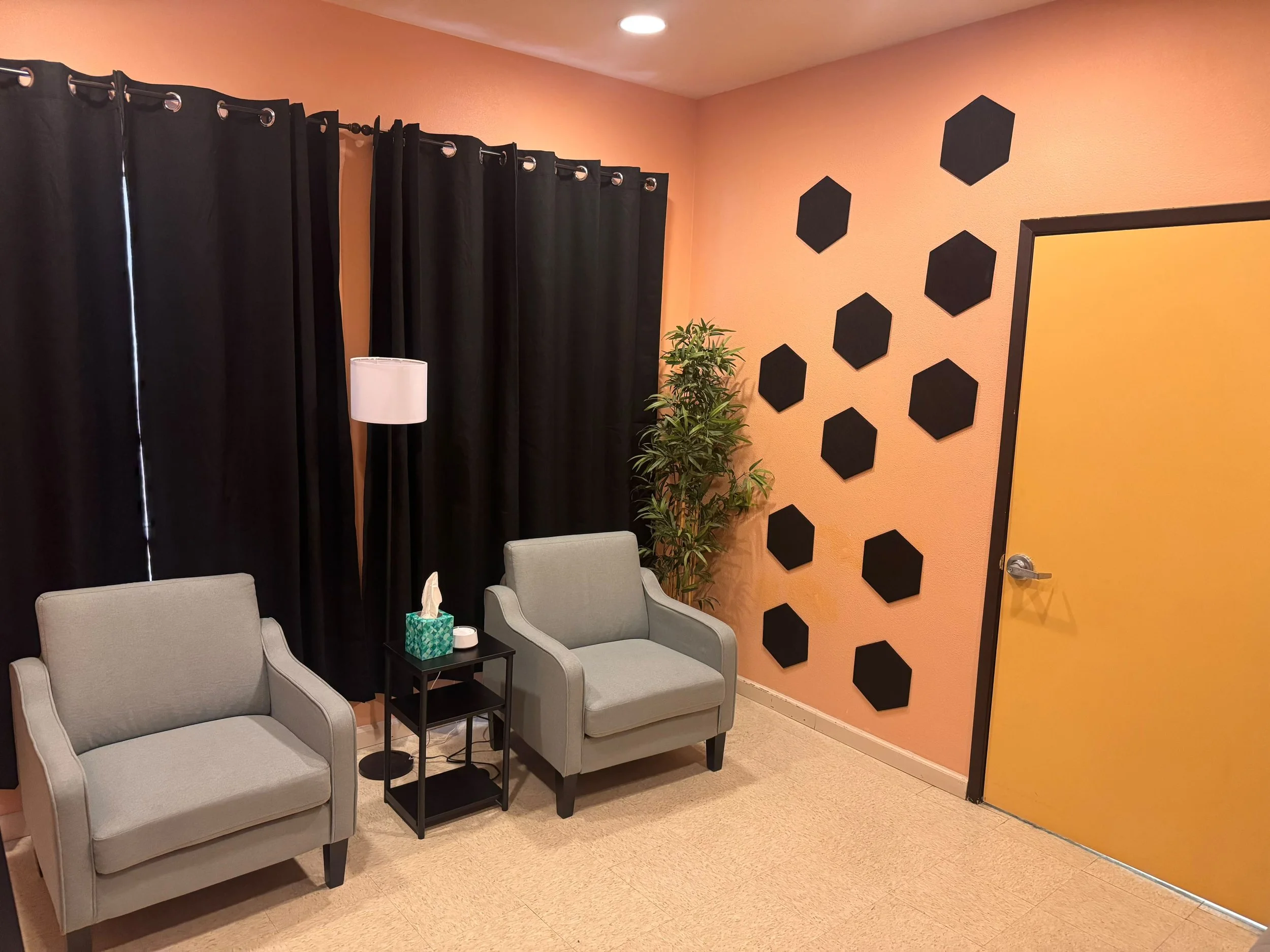 A waiting room with two light gray armchairs, a black side table holding tissues and sanitizer, a lamp, and a potted plant. The wall is painted orange with black hexagonal decoration, and there are black curtains covering a window.