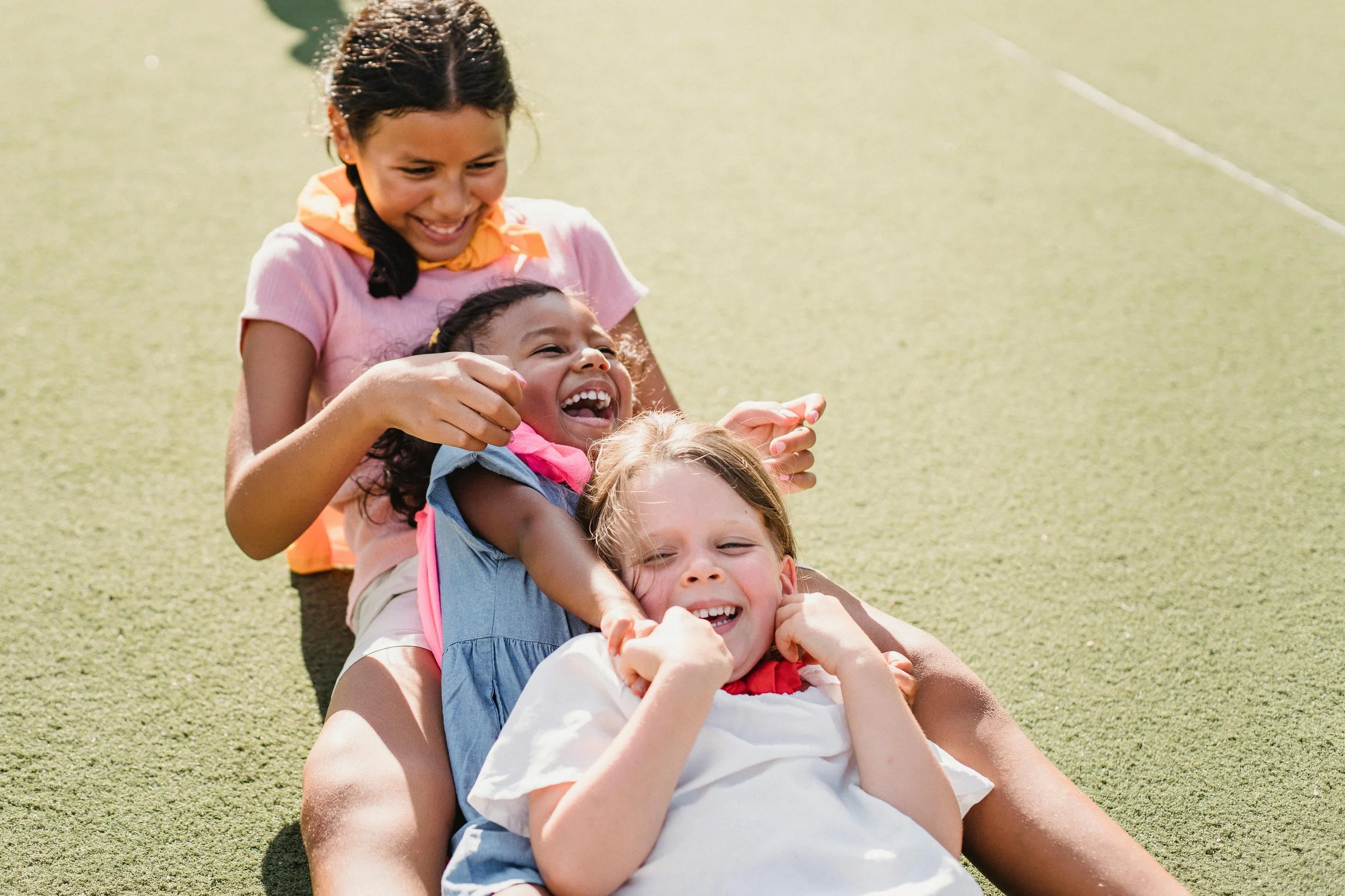 Four children sitting on the ground, laying back to back, laughing and playing on a grassy outdoor field.