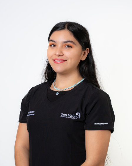 Portrait of a young woman with long dark hair wearing a black shirt and a beaded necklace, smiling at the camera against a plain white background.