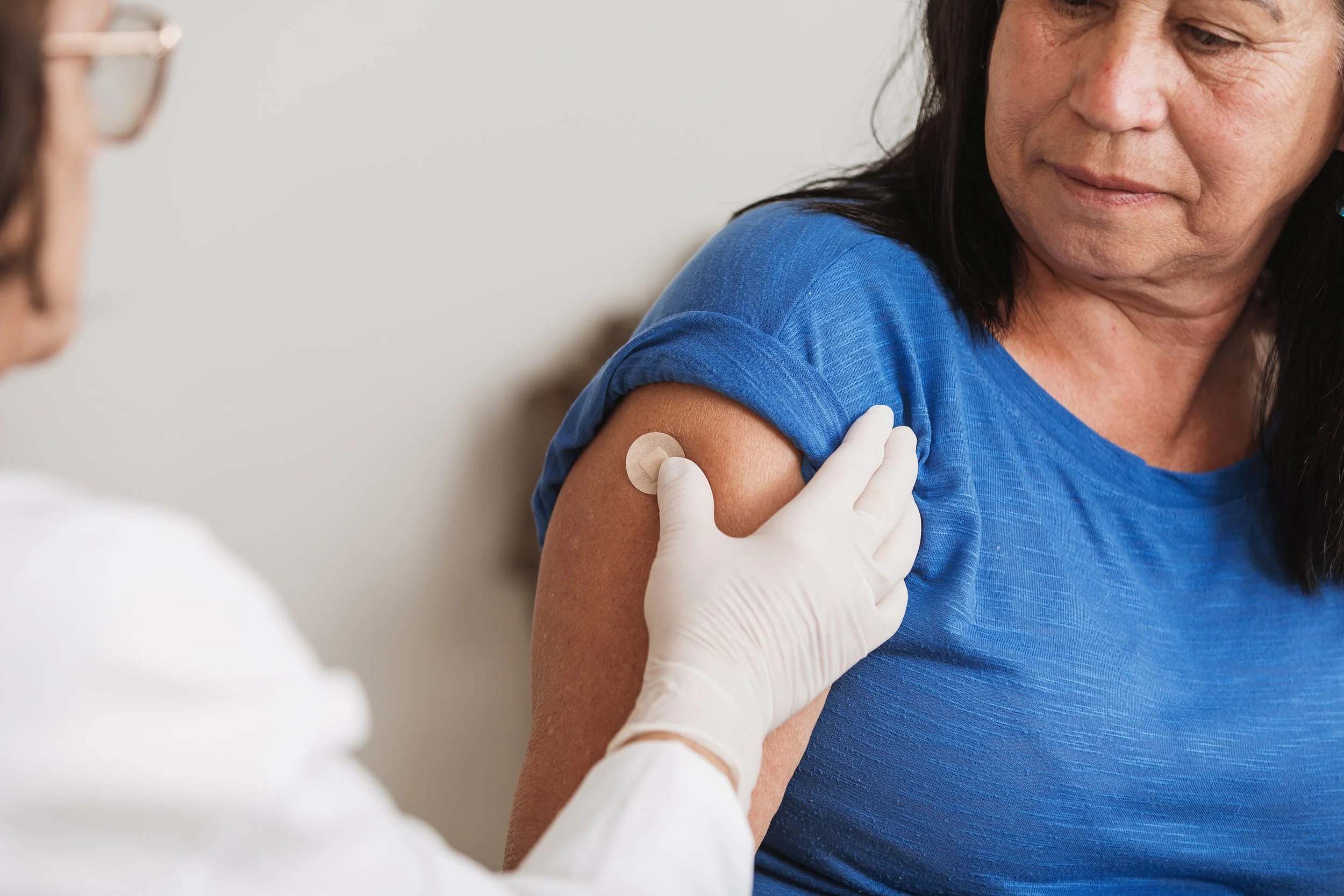 A woman receiving a vaccine injection in her upper arm from a healthcare professional wearing white gloves.