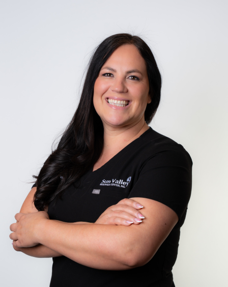 A smiling woman with long dark hair, wearing a black shirt with a logo, standing with her arms crossed against a plain white background.