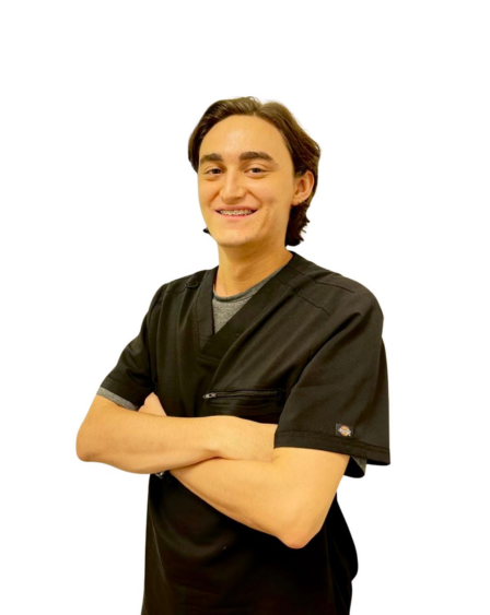 Young man with dark hair, wearing medical scrubs, smiling with arms crossed, standing against a white background.