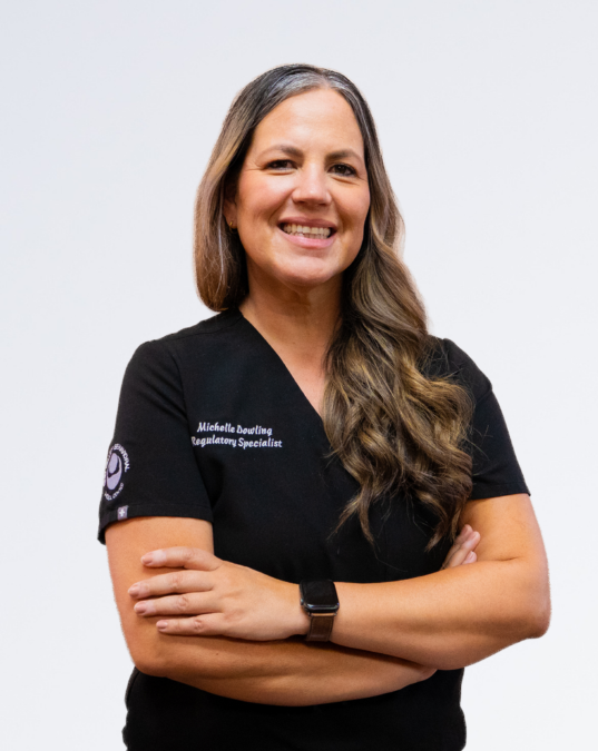 A woman with long wavy brown hair, wearing a black polo shirt, standing with arms crossed and smiling against a plain white background.