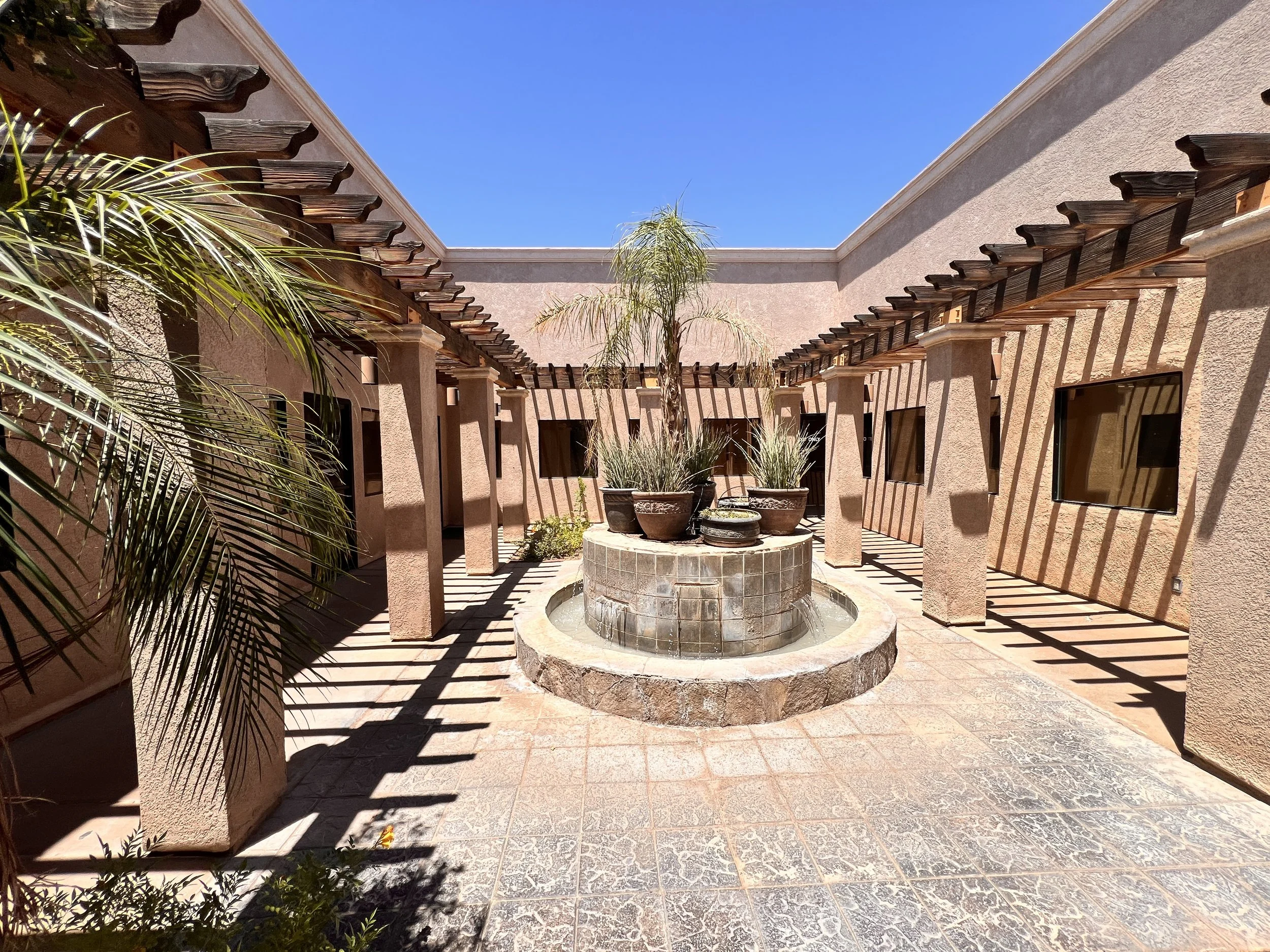 Courtyard with a fountain surrounded by potted plants, stucco walls, and wooden pergola beams, under a clear blue sky.
