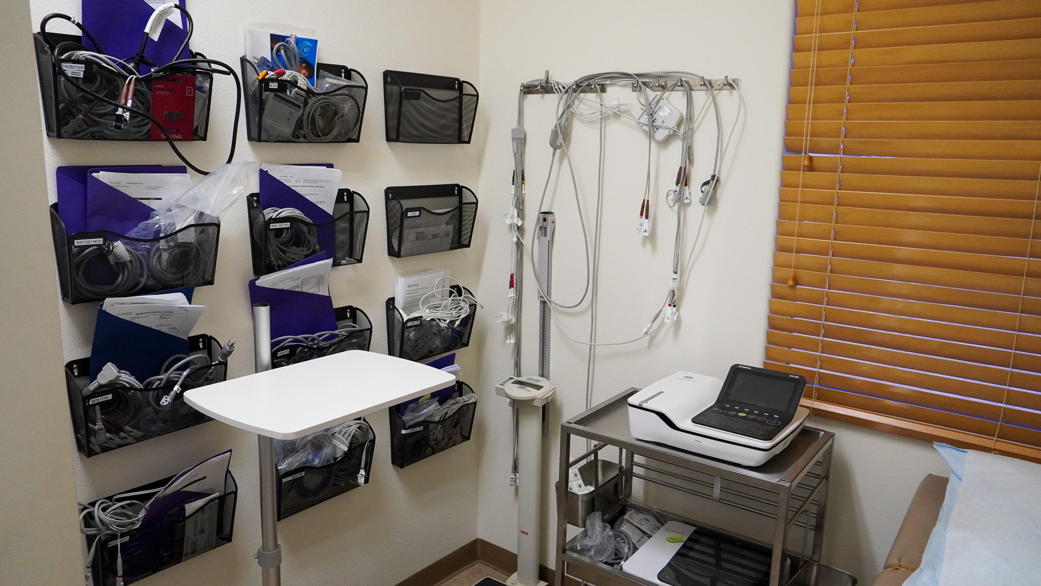 Medical or office examination room with wall-mounted black mesh bins filled with cables, papers, and tools on the left wall, medical equipment hanging on a wall hook in the corner, a window with wooden blinds on the right, and a white tabletop, scann