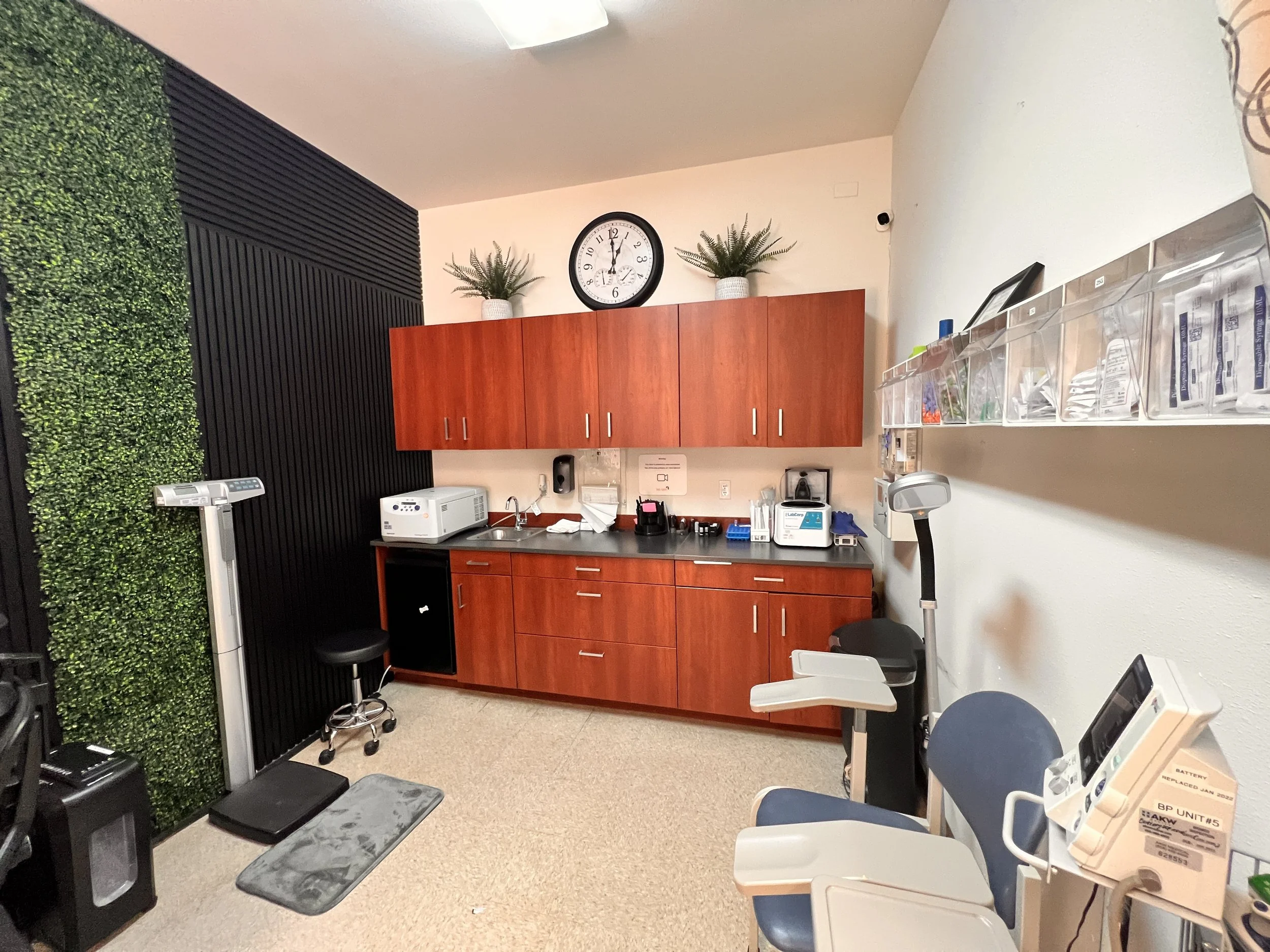 Interior view of a medical or clinical room with a wall-mounted clock showing 12:02, two potted plants on top of wooden cabinets, and various medical equipment and supplies on countertops and shelves. There is a green leafy wall decoration on the left and a black panel on the far left side.