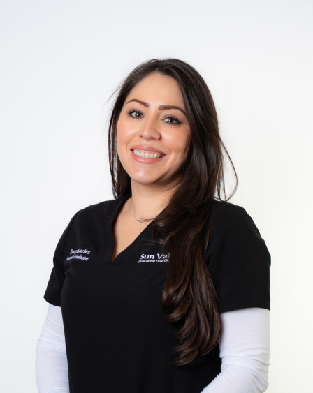 A smiling woman with long dark hair wearing a black medical uniform and a white long-sleeve shirt underneath, standing against a white background.