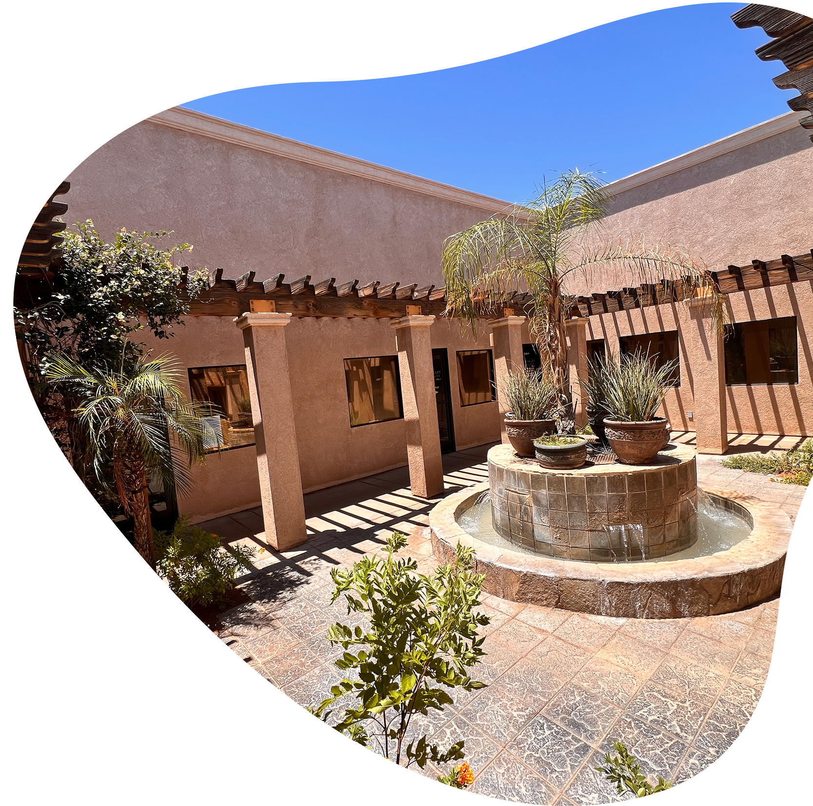 A courtyard with a small circular water fountain at the center, surrounded by potted plants, a wooden pergola, and beige stucco walls with several windows, under a clear blue sky.