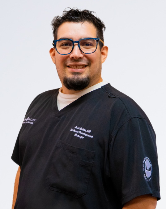 A man with dark curly hair, glasses, and a goatee, wearing a black medical scrub top with white lettering and a logo on the sleeve, smiling against a plain white background.