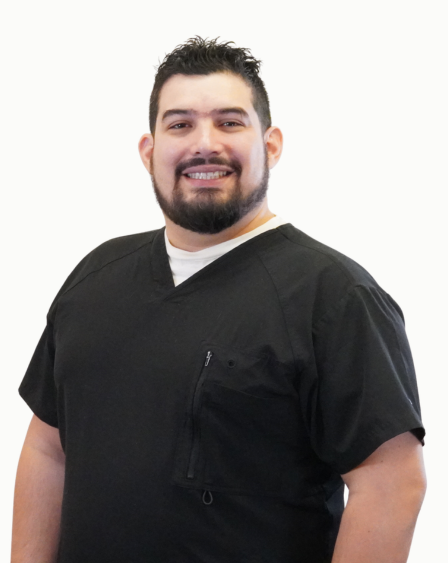 A smiling man with dark hair and a beard, wearing a black medical scrub top, standing against a plain white background.